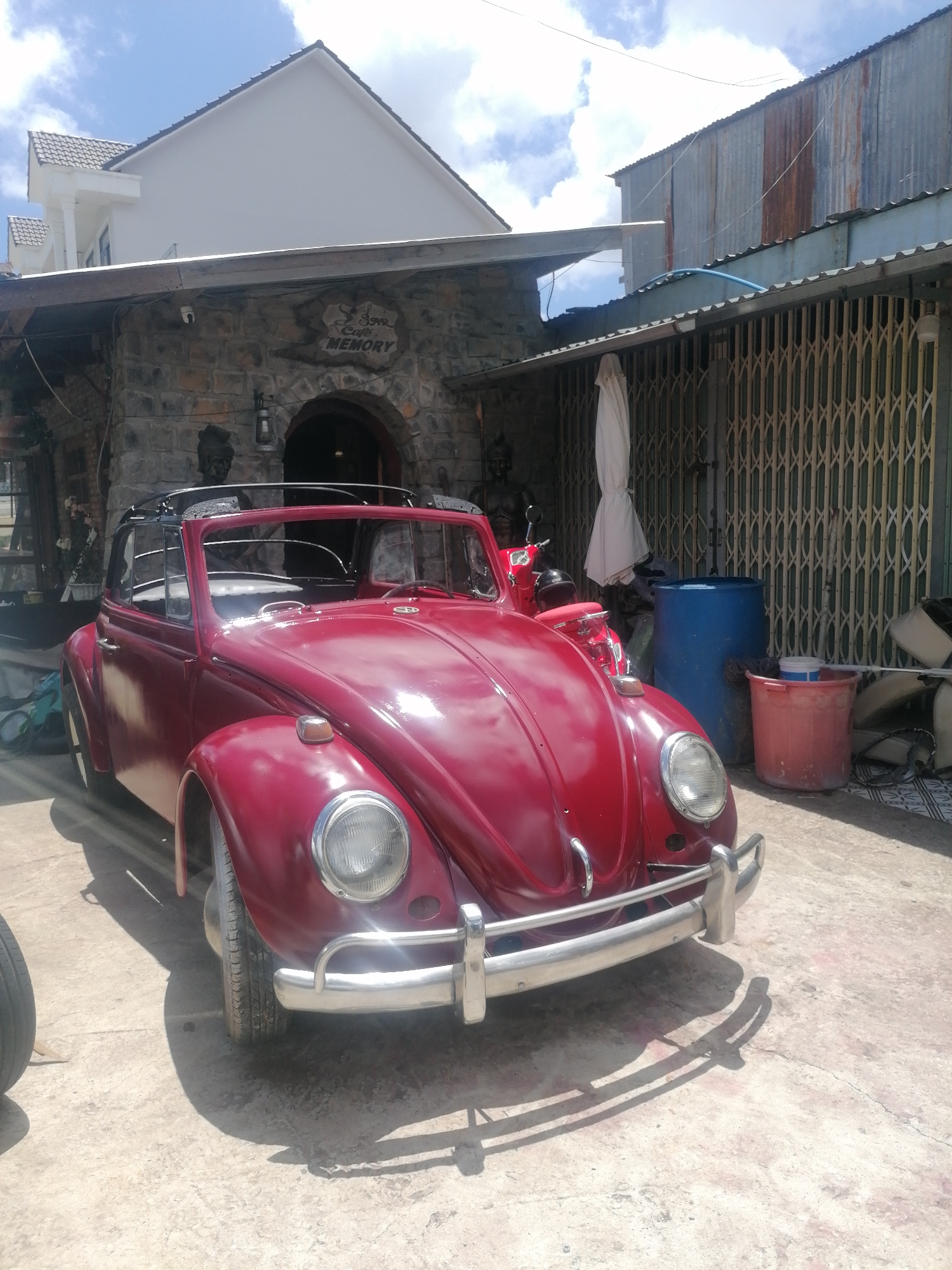 A front, three-quarter view of a shiny, maroon convertible Volkswagen Beetle is parked on a concrete surface. The car is positioned at a slight angle, with its front bumper pointing towards the viewer. The convertible top is down, revealing the interior. The car has large, circular headlights and a chrome bumper.
Behind the car, a stone building with an arched entryway is visible. Above the archway, a sign reads "Cafe Memory". To the right of the stone building, there is a corrugated metal structure with a rust patina. In front of this structure, a large blue barrel and a red bucket are placed on the ground. White fabric, possibly an umbrella or a drape, hangs from the corrugated metal roof.
The scene appears to be outdoors, with a bright sky and scattered clouds overhead. Sunlight casts shadows on the ground, indicating a sunny day. The overall impression is that of a vintage car parked outside a cafe or a shop in Da Lat, Vietnam, a city known for its French colonial architecture and scenic beauty.