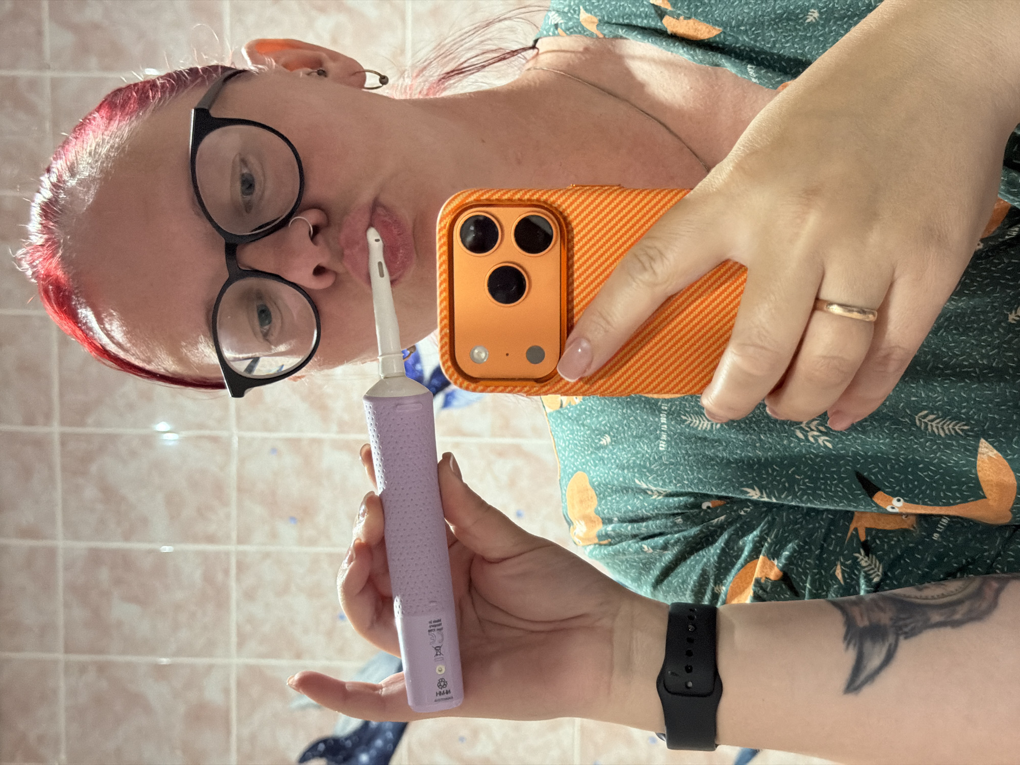A woman is taking a selfie in a bathroom. She has red hair and is wearing glasses. She is holding a purple electric toothbrush in one hand and an orange smartphone in the other. The phone case has a textured pattern and three camera lenses. The woman's skin is fair, and she has a wedding ring on her finger. A black smartwatch is visible on her wrist, and a tattoo is present on her forearm. She is wearing a green top or pajamas with a fox pattern. The bathroom has light pink tiles. There is no indication that the location is Chaikovsky, Russia.