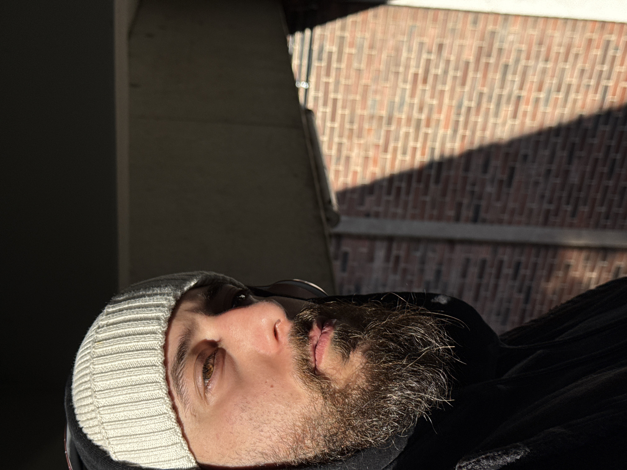 A male individual is depicted in the foreground of an outdoor scene in Strasbourg, France. He possesses light skin, brown eyes, and a prominent dark brown beard and mustache. He is wearing a light cream-colored, ribbed knit beanie and a dark, presumably black, hooded garment with the hood partially visible. Strong, direct sunlight illuminates the right side of his face, including his eye, nose, and cheekbone, while the left side of his face and body remains in shadow. A thin, dark object is faintly visible near his right ear, consistent with an earphone or headphone frame. The subject's gaze is directed slightly upward and to his right. The background features a dark, flat concrete-like overhead structure, casting a distinct shadow. Below and to the right, a reddish-brown brick wall, constructed in a stretcher bond pattern, is partially visible, also marked by a strong diagonal shadow from the direct sunlight. No other individuals or significant objects are clearly identifiable.