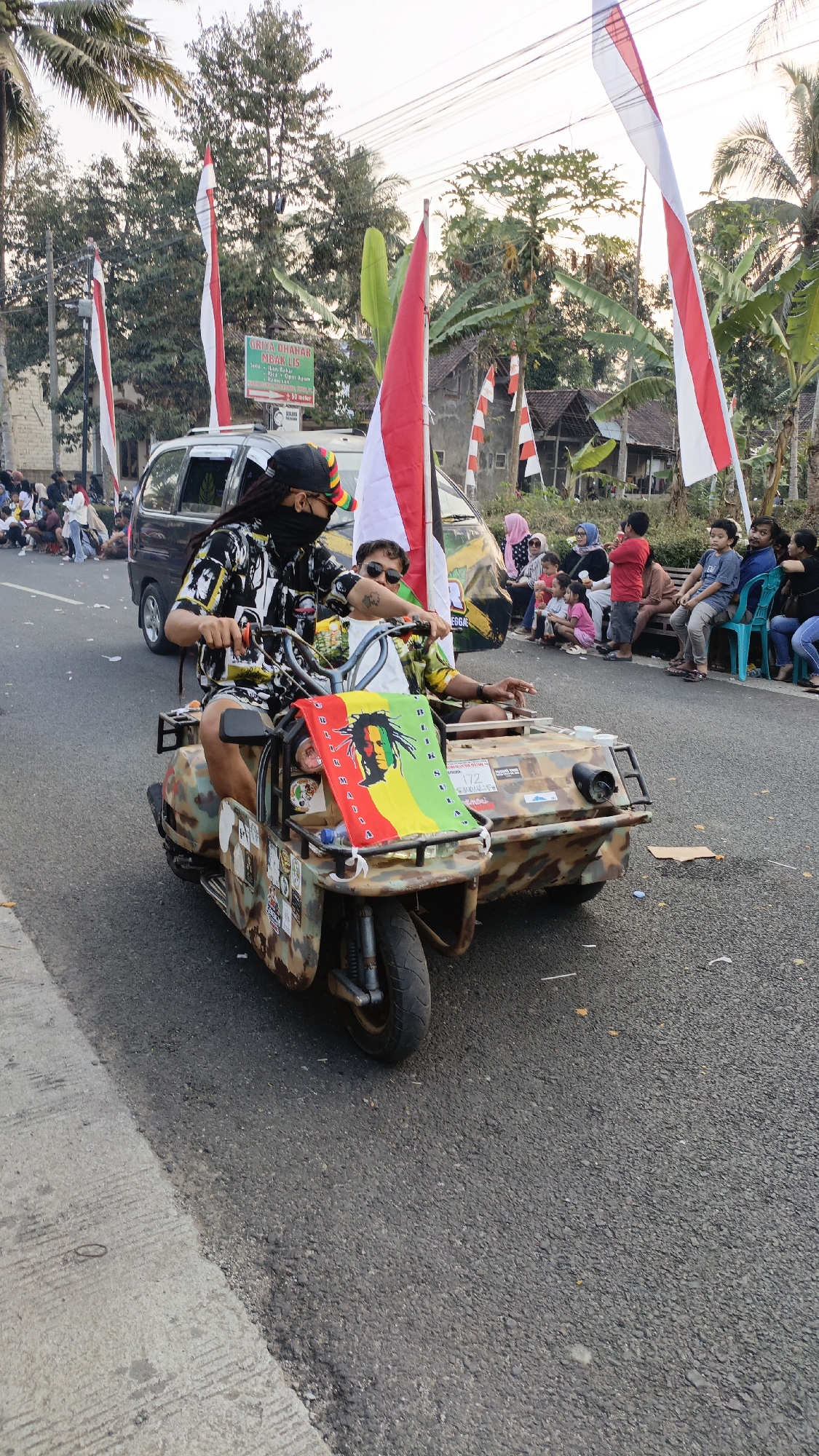In Krajan Lor, Indonesia, two individuals ride a three-wheeled vehicle modified with a sidecar, decorated with a Rastafarian flag. The driver, wearing a black bandana, black and white patterned shirt, and a black cap with colorful stripes, steers the vehicle. The passenger, wearing sunglasses and a patterned shirt, sits in the sidecar. The vehicle itself is camouflaged with a brown, green, and black pattern and displays various stickers. Red and white Indonesian flags are prominent, flying from poles attached to the vehicle and from poles along the roadside. Spectators are gathered on the side of the road, some seated on benches and others standing, observing the procession. A van is visible behind the three-wheeled vehicle, also adorned with flags. The scene takes place on a paved road lined with palm trees and houses, suggesting a street in a village or town during a festive event or parade.