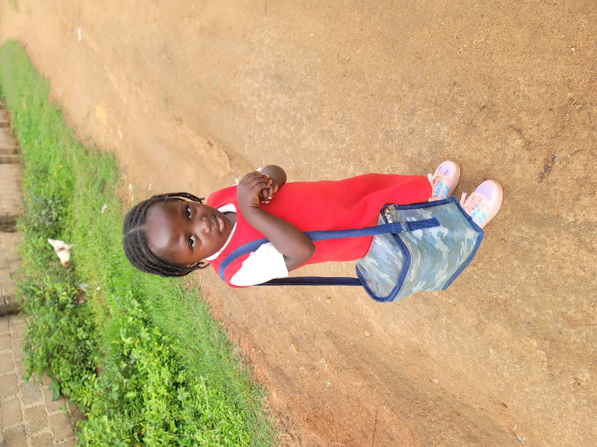 A young Black girl is standing in a dirt path next to a patch of green grass. She is wearing a red dress with a white shirt underneath, and light pink sneakers with blue accents. She has her hair braided in pigtails. A camouflage patterned bag with blue trim is slung over her shoulder. She is looking directly at the camera with a neutral expression. To the left of the path, there is a brick wall and thick green foliage with some yellow flowers. In the grass, a white chicken is visible. The scene appears to be outdoors during the daytime, with natural lighting. The dirt path suggests a rural or unpaved area.