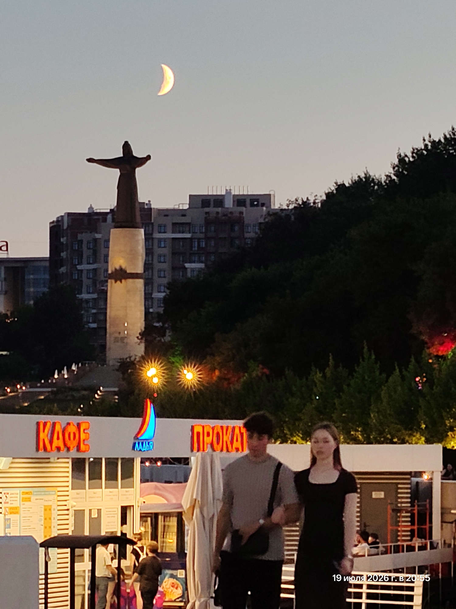 An outdoor, nighttime scene in an unknown city shows a bronze statue of a figure with outstretched arms on a tall pedestal, illuminated by spotlights. Above the statue, a crescent moon is visible in the pale sky. In the foreground, two young people, a male and a female, walk arm-in-arm, facing the viewer. They are dressed in casual attire. Behind them, a building with bright neon signs reading "КАФЕ" and "ПРОКАТ" is visible. The surrounding area appears to be a public space with trees, lights, and other structures suggesting a waterfront or entertainment district. A date and time stamp, "19 июля 2026 г. в 20:55", is superimposed on the lower right corner of the image.