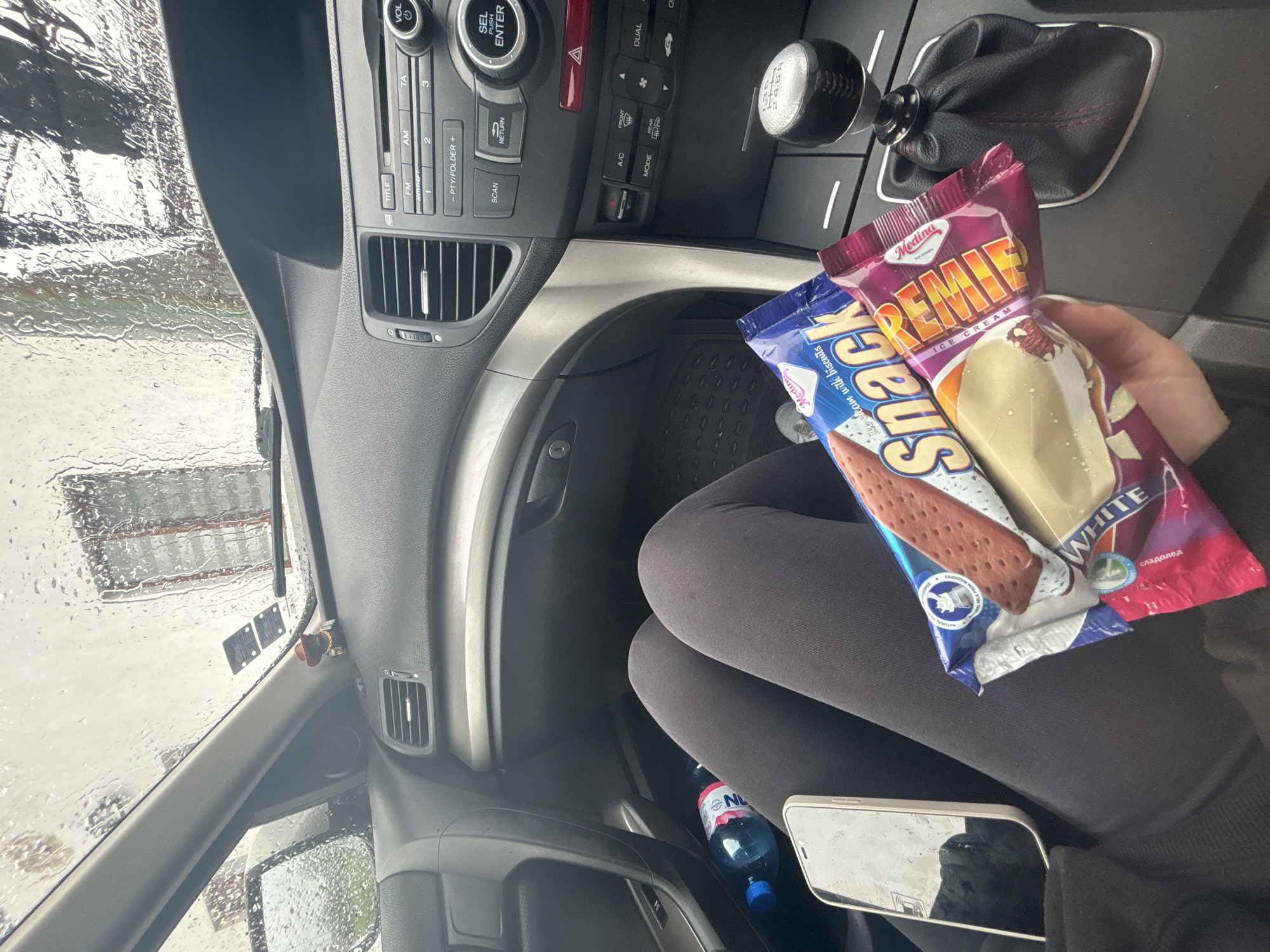 This media file captures the interior of a car from a high angle, looking towards the front and right. A person, wearing dark leggings, is seated, holding two packages of ice cream. The blue package is labeled "Medina Ice cream with biscuits Snack" and the pink/purple one says "Medina PREMIE ICE CREAM," featuring "WHITE" and the Bulgarian word "сладолед" (ice cream).
The car's dashboard and center console are prominent, displaying an audio system with buttons for "VOL", "SEL PUSH ENTER", "FM", "AM", "TA", "TITLE", "SCAN", "RETURN", and "PTY/FOLDER +". Climate control buttons for "A/C", "DUAL", and "MODE" are also visible, alongside a hazard light switch. A manual gear shifter with a black leather boot and red stitching shows a "6 R" pattern.
The windshield and side mirror are covered in raindrops, indicating wet weather conditions, suggesting the car is either in the rain or a car wash. A water bottle with a "NL NATURAL" label and a smartphone are resting on the floor beside the person's leg. The scene takes place during the daytime, and the text "сладолед" aligns with the location context of Bulgaria.
