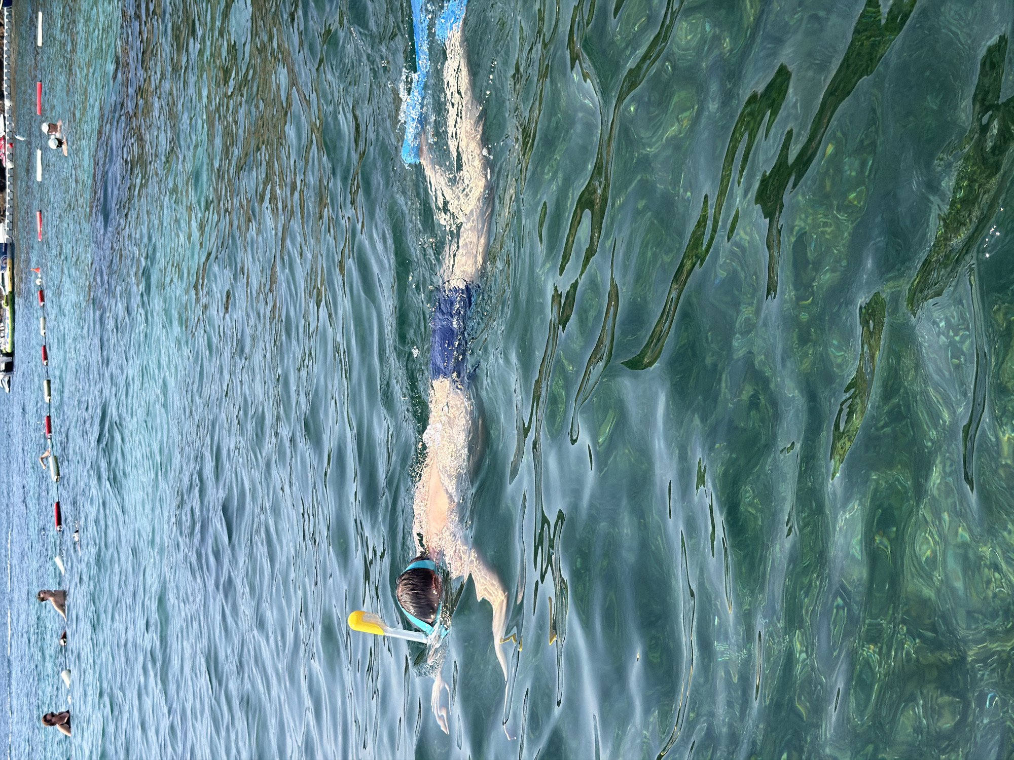 In the clear, turquoise waters of Budva, Montenegro, a person is snorkeling. They are wearing a blue mask with a yellow snorkel, blue swim trunks, and blue fins. The water is calm, with visible ripples on the surface, and sunlight refracts through the water, illuminating the sandy bottom. In the background, other swimmers are visible in the distance, near a line of buoys. The scene suggests a leisurely day at the beach.