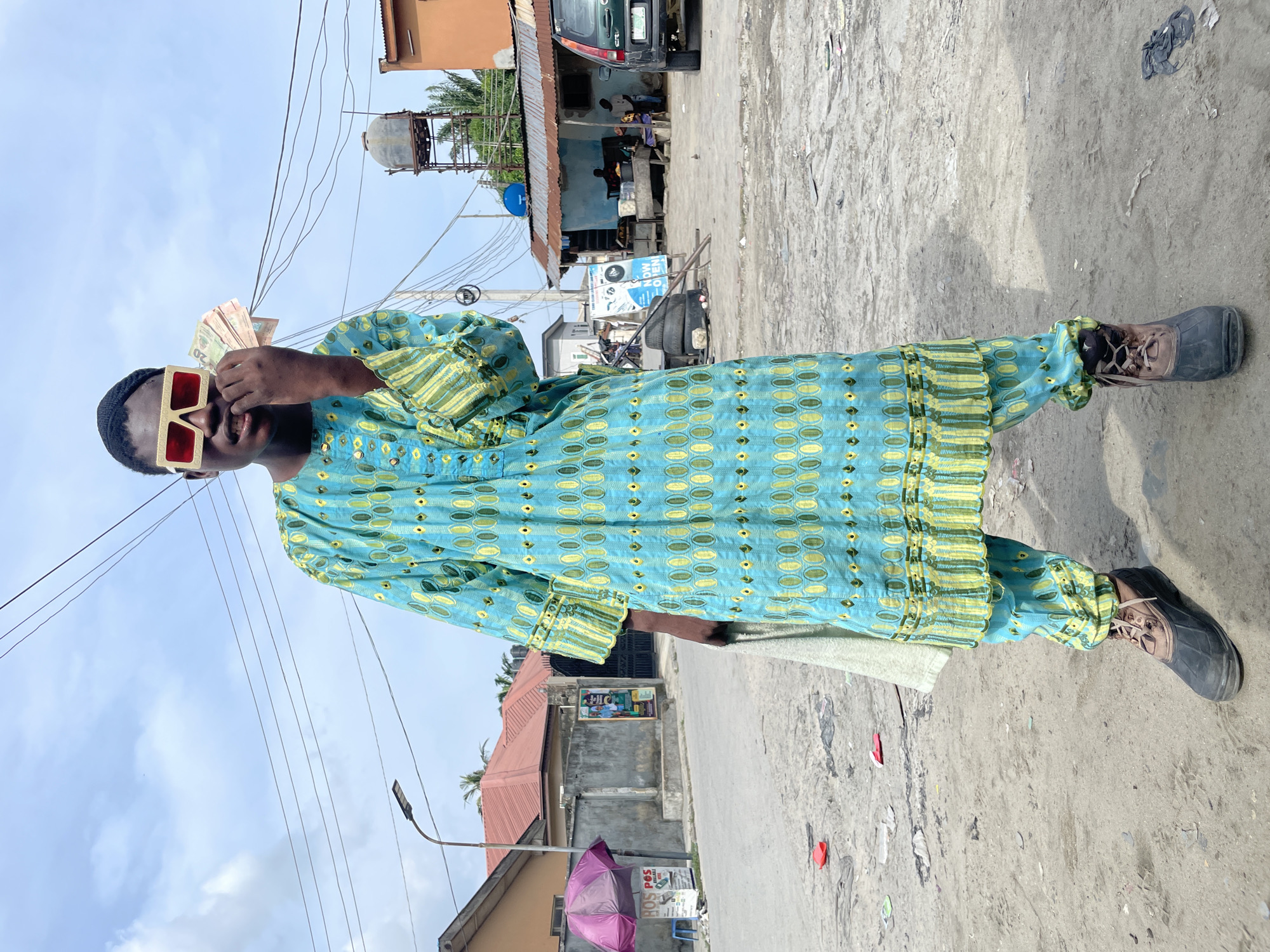 A man wearing an aqua and yellow patterned shirt and matching pants stands on a dirt and concrete road in Ado, Nigeria. He wears large, red-framed sunglasses and holds a wad of cash in front of his face. The road is littered with trash and debris. Buildings and structures surround the road, including a blue building with people underneath a corrugated metal roof, utility poles with wires, and houses. An overhead water storage tank is visible in the background.