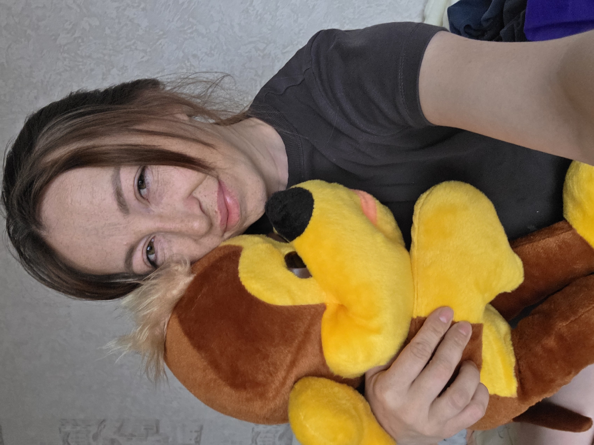 A woman is lying down, holding a stuffed animal, possibly indoors. The woman has brown hair with highlights and freckles. She is wearing a dark grey t-shirt. The stuffed animal is brown and yellow with a black nose. The location is Unknown, Unknown.