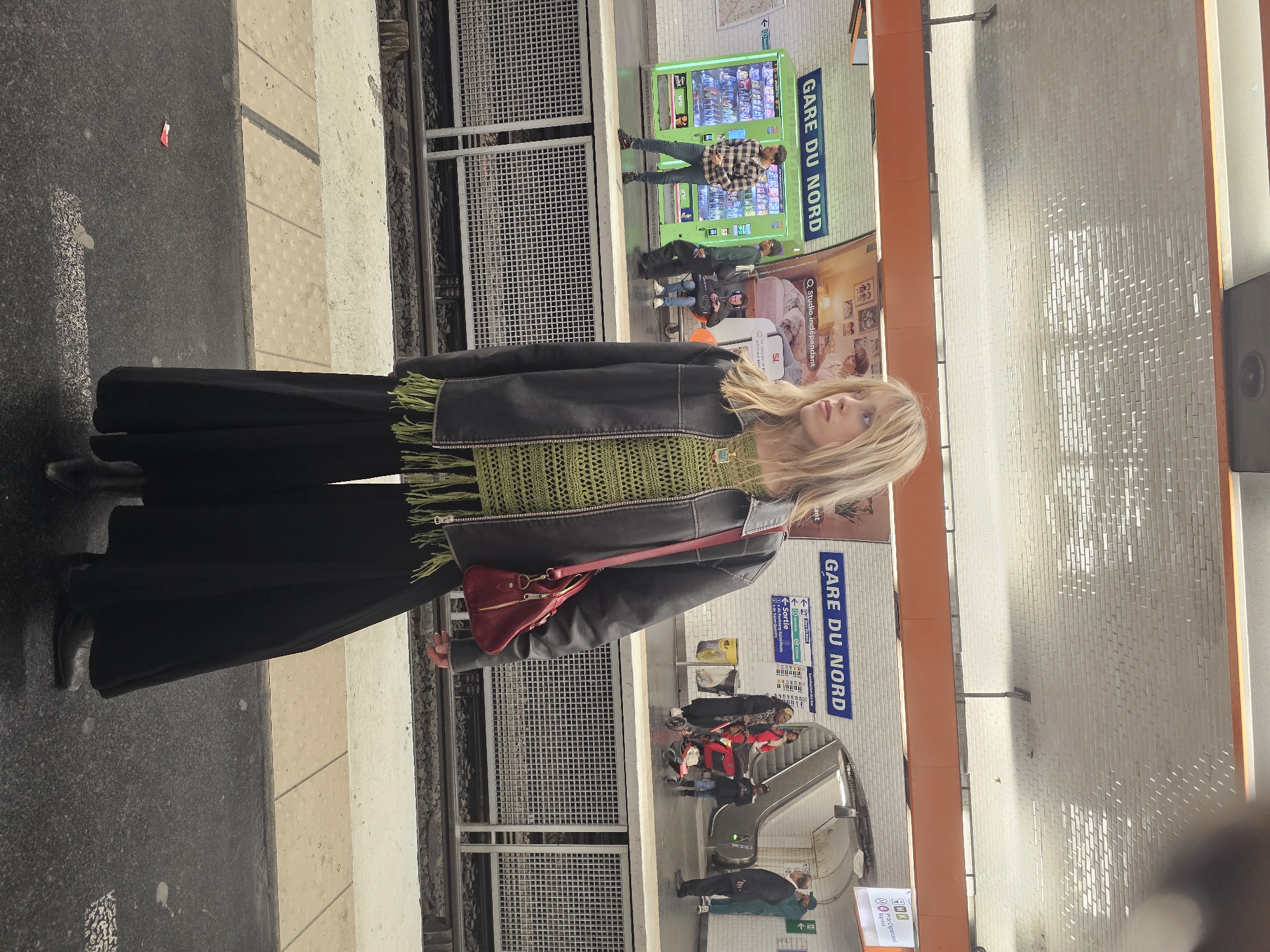 An image captures a young woman standing on a subway platform at Paris's Gare du Nord station. The woman, with long blonde hair, is the central subject, dressed in a black jacket layered over a green knitted top with fringed details, paired with wide-leg black trousers and dark shoes. A small red shoulder bag is slung across her body. She is looking slightly upwards and to her right.
The setting is a well-lit subway platform. In the background, across the tracks, another platform is visible with several people. A man in a plaid shirt stands near a prominent bright green vending machine. Further back, an escalator leads down, with a few individuals, including someone pushing a stroller, nearby. White tiled walls line the station, and metal grilles separate the tracks from the platform edges.
The activity appears to be waiting for a train or transiting through the station. The overhead lighting suggests a bright indoor environment, characteristic of a metropolitan subway system.
Notable details include multiple signs confirming the location: "GARE DU NORD" is visible on blue signs in two places. Other directional signs indicate "← Sortie" (Exit) and specific street names like "du Fasbourg Saint-Denis" and "21.de Saint-Quentin", providing further geographical context within Paris. Additional text, "Q Studio indépendant," "AHK," and "Bagtest," can also be seen on various posters or displays. A small red item, possibly litter, is visible on the platform closer to the tracks.