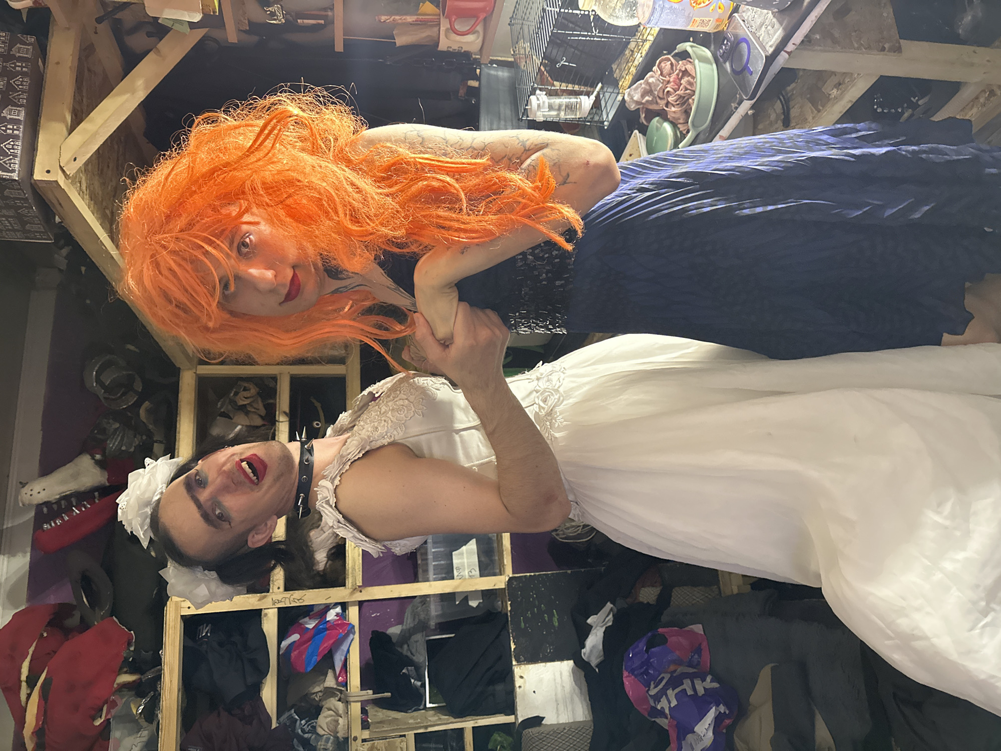 The image depicts two individuals in a cluttered indoor environment, possibly a workshop or storage area. One individual, on the left, is wearing a white wedding dress and a black spiked collar. They have dark hair styled with a white bow and are looking towards the camera with makeup applied. The second individual, on the right, has bright orange, voluminous hair and is wearing a dark, sequined top and a blue, striped skirt. They are holding hands with the first individual and looking towards the camera with makeup applied. The background is filled with shelves containing various objects, including clothing, containers, and what appear to be props or costumes. A cage and shelves with items are visible on the right side of the image. The overall scene is dimly lit. The provided image does not contain any information or visual cues that would allow for the identification of its location in the city of Saratov, Russia.
