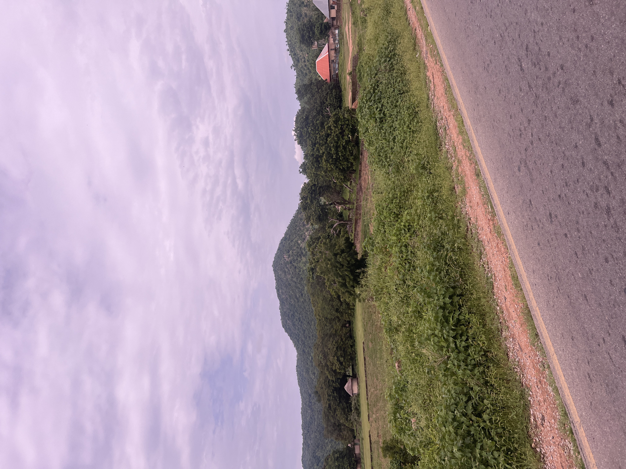 The image depicts a roadside scene in Minda, Nigeria. A paved road runs diagonally from the upper right to the lower left of the frame, bordered by a narrow strip of sandy earth and then dense, green vegetation. Beyond the vegetation, a rolling, tree-covered hill rises, its slopes meeting a softly clouded sky. In the mid-ground, nestled among the trees, are a few buildings. One building features a prominent, bright orange-red roof. In the far distance, the hill continues to recede. The lighting suggests it is daytime, likely overcast or late afternoon, given the soft, diffused light and the purple hue of the sky. The scene conveys a sense of rural tranquility, with the natural landscape dominating the view. There is no visible text in the image.