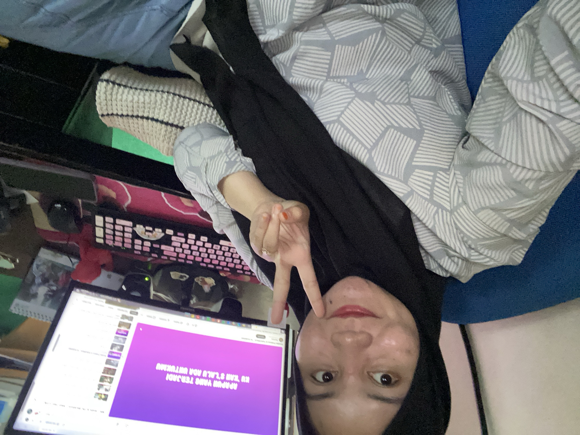 A woman wearing a black hijab and a grey and white patterned shirt is seated in a blue chair, holding up two fingers in a peace sign. She is looking at the camera with a slight smile. Behind her is a computer monitor displaying text in Indonesian on a purple background. A pink and black keyboard is in front of the monitor. The scene appears to be indoors, possibly in a home office or study in Banjarbaru, Indonesia.