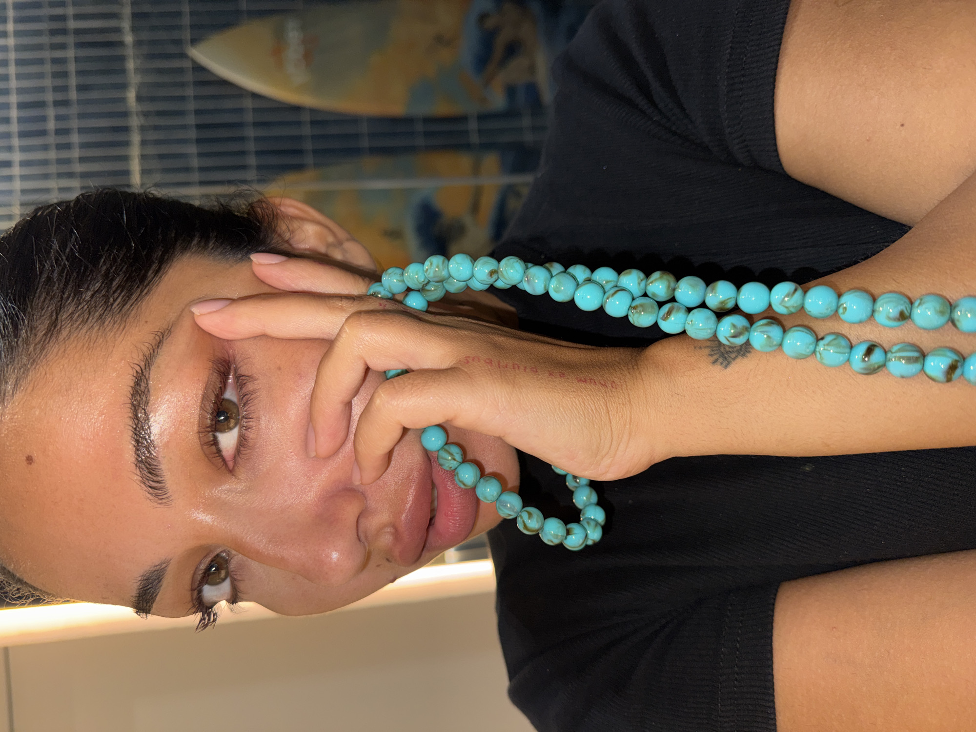 A woman with brown eyes is holding a strand of turquoise beads in her right hand. She is wearing a black shirt. There is a surfboard visible in the background, and the location is Kuta Selatan, Indonesia.