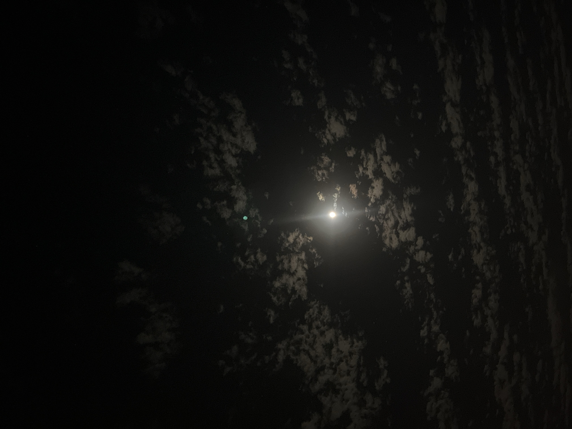 The image depicts a night scene in an unknown city. A bright moon shines through scattered clouds, casting light on some of them. The majority of the image is dark.
