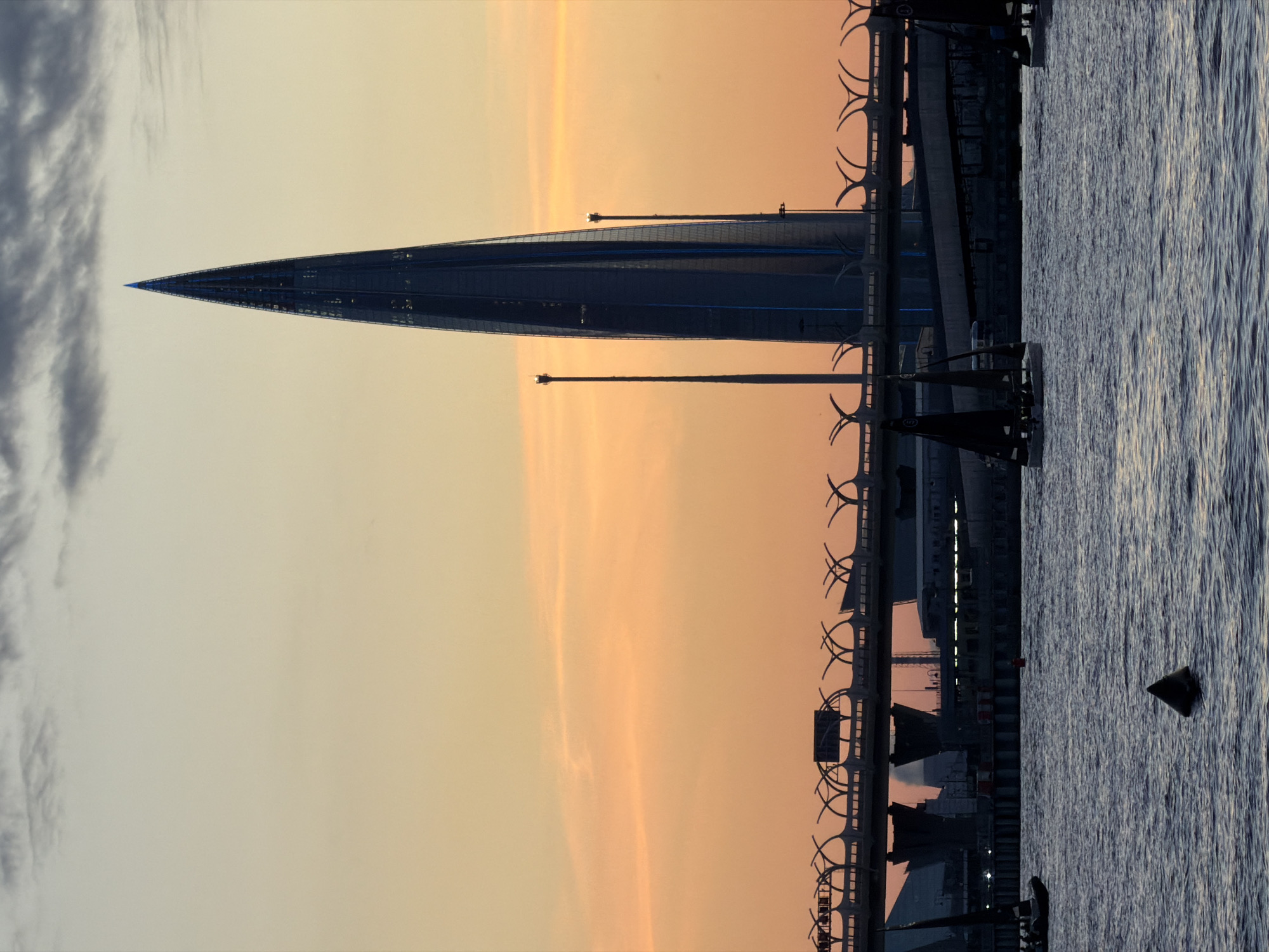 The image depicts a serene water scene in Saint Petersburg, Russia, during the golden hour of sunset. Dominating the skyline is the iconic Lakhta Center, a futuristic skyscraper with a pointed, spire-like design, illuminated with subtle blue lights. In the foreground, several sailboats with dark sails glide across the rippling water. Behind them, a modern bridge with its distinctive structural elements spans the waterway. The sky is a gradient of warm orange and soft yellow, with wisps of grey clouds adding texture. The water reflects the warm hues of the sky and the silhouettes of the structures. A lone triangular buoy floats near the center of the frame, adding a subtle navigational marker to the tranquil maritime scene.