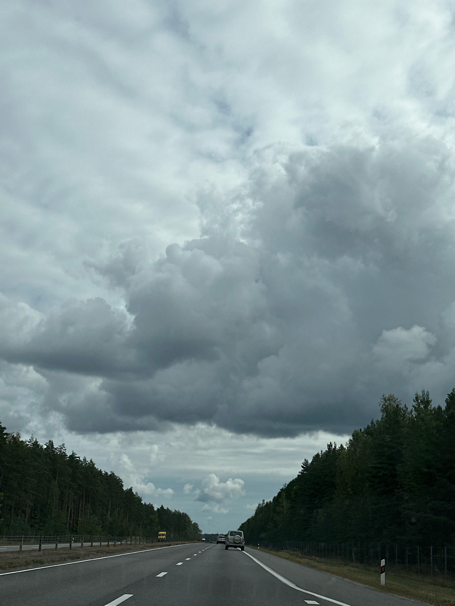 The image depicts a highway scene in the Вяззеўскі сельскі Савет, Belarus, under a cloudy sky. The road is asphalt with clear white lane markings. On the left side of the road, a dense forest of green coniferous trees lines the highway, separated by a fence. On the right side, the forest also stretches along the road. Several vehicles are visible on the highway, including a white SUV in the foreground on the right lane and other cars further ahead. A yellow truck is seen on the left side of the road in the distance. The overall atmosphere is overcast with heavy, dark clouds dominating the sky.
