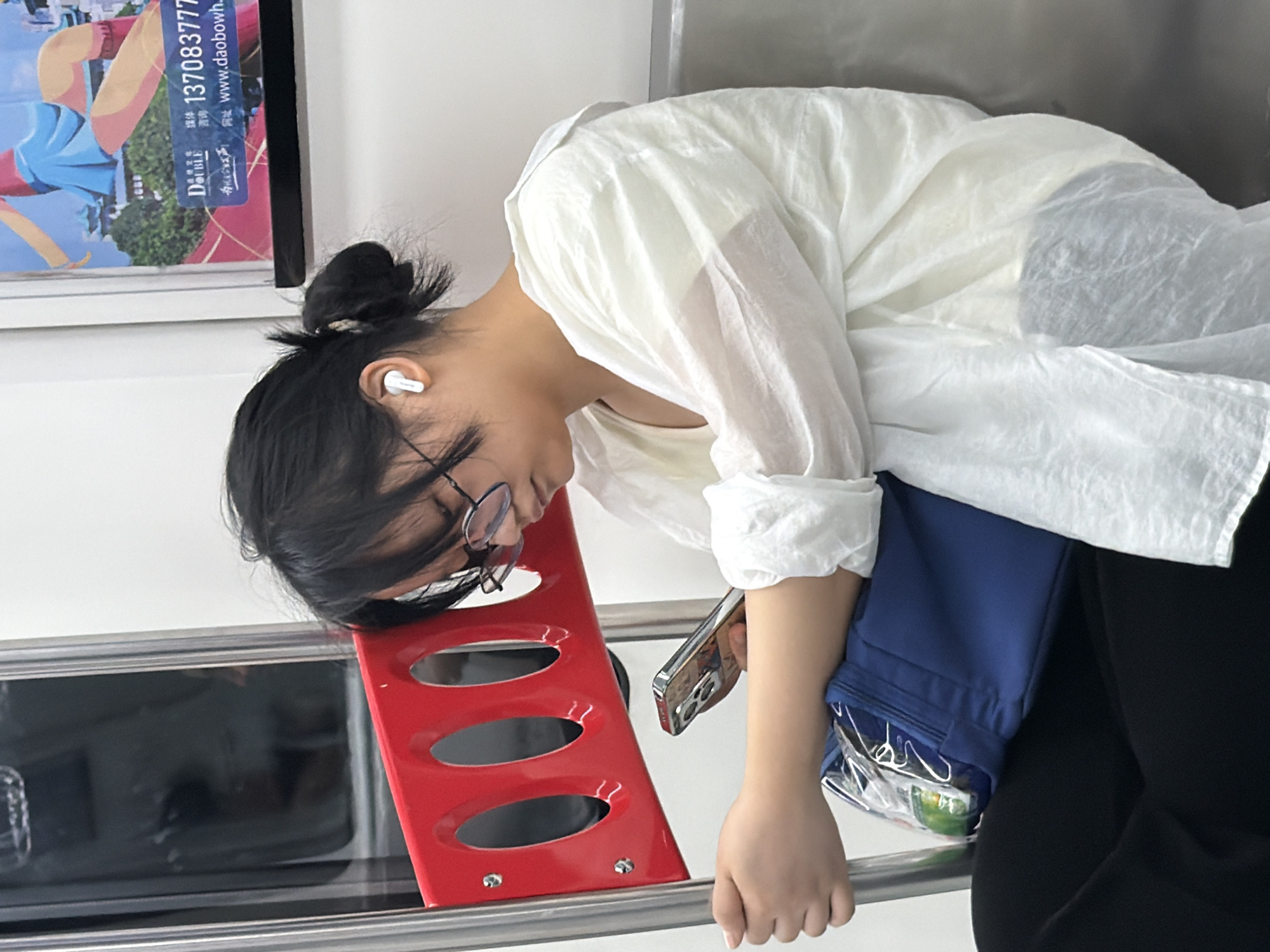 A young East Asian female is seated on public transportation, likely a train or subway, in Yuzhong District, China. She wears black-rimmed glasses, a white loose-fitting shirt, and black pants. Her dark hair is pulled back into a bun, with some strands loose around her face. She has white wireless earbuds in her ears. In her hands, she is holding a smartphone, looking down at it. To her left, a blue bag is partially visible on her lap, containing what appears to be packaged snacks. Behind her, a red metal bar with oval cutouts is attached to the wall. A poster with an animated figure and Chinese text is affixed to the wall above her. The environment appears to be the interior of a transit vehicle, with a gray seat and metal support poles visible.