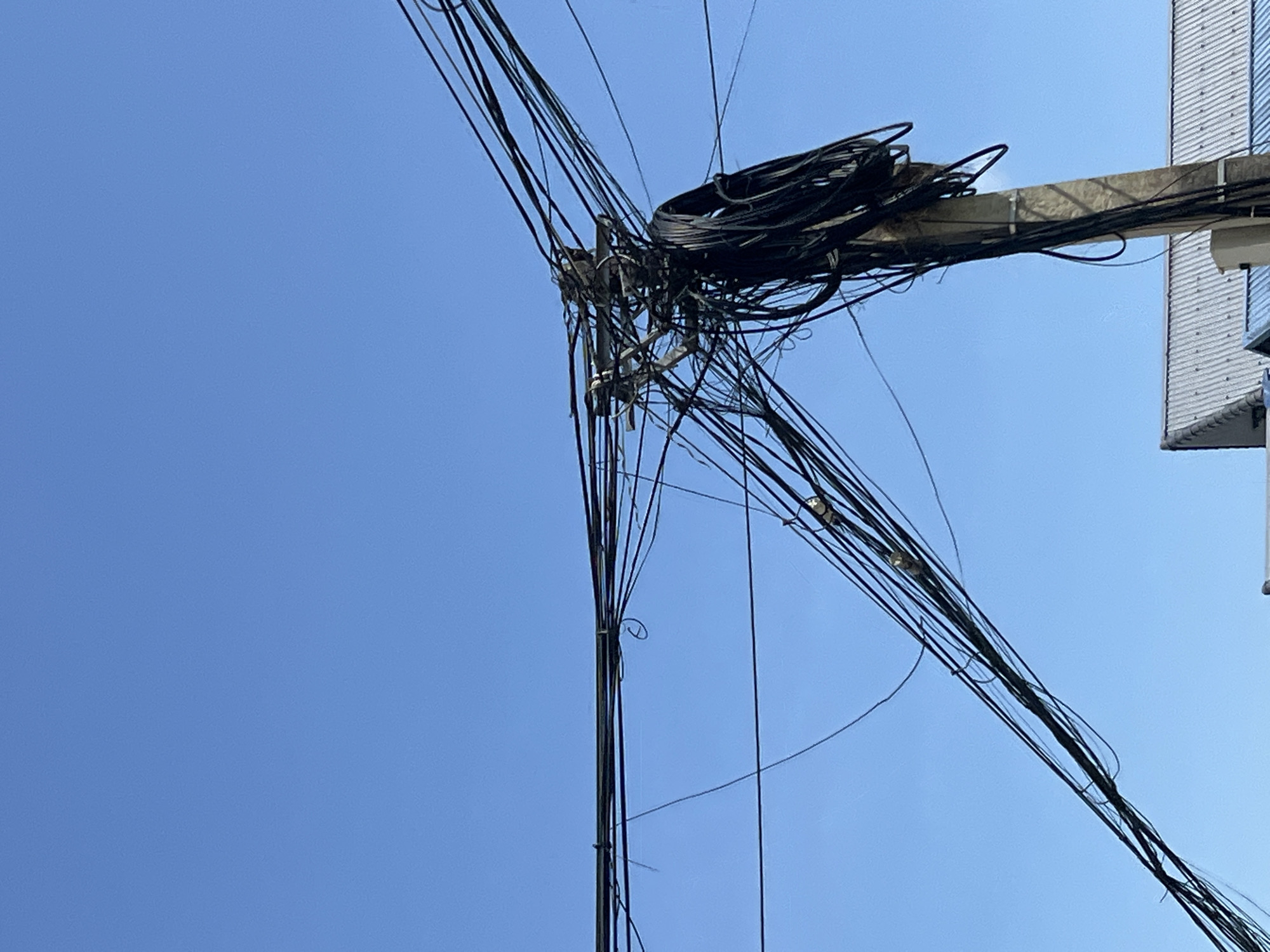 An outdoor, low-angle, eye-level medium shot shows a utility pole laden with a dense cluster of black wires against a clear, pale blue sky. The pole, made of concrete, is positioned in the lower right quadrant of the frame, extending upwards and disappearing into the mass of tangled wires. The wires, varying in thickness and length, crisscross and loop around each other, creating a chaotic and complex network. Some wires hang loose, while others are tightly bound together. In the lower portion of the image, the corrugated metal roof of a building is visible beneath the utility pole, suggesting an urban or semi-urban setting. The scene is devoid of any people or animals. The overall impression is one of untamed electrical infrastructure in the town of Bản Bo, Vietnam.