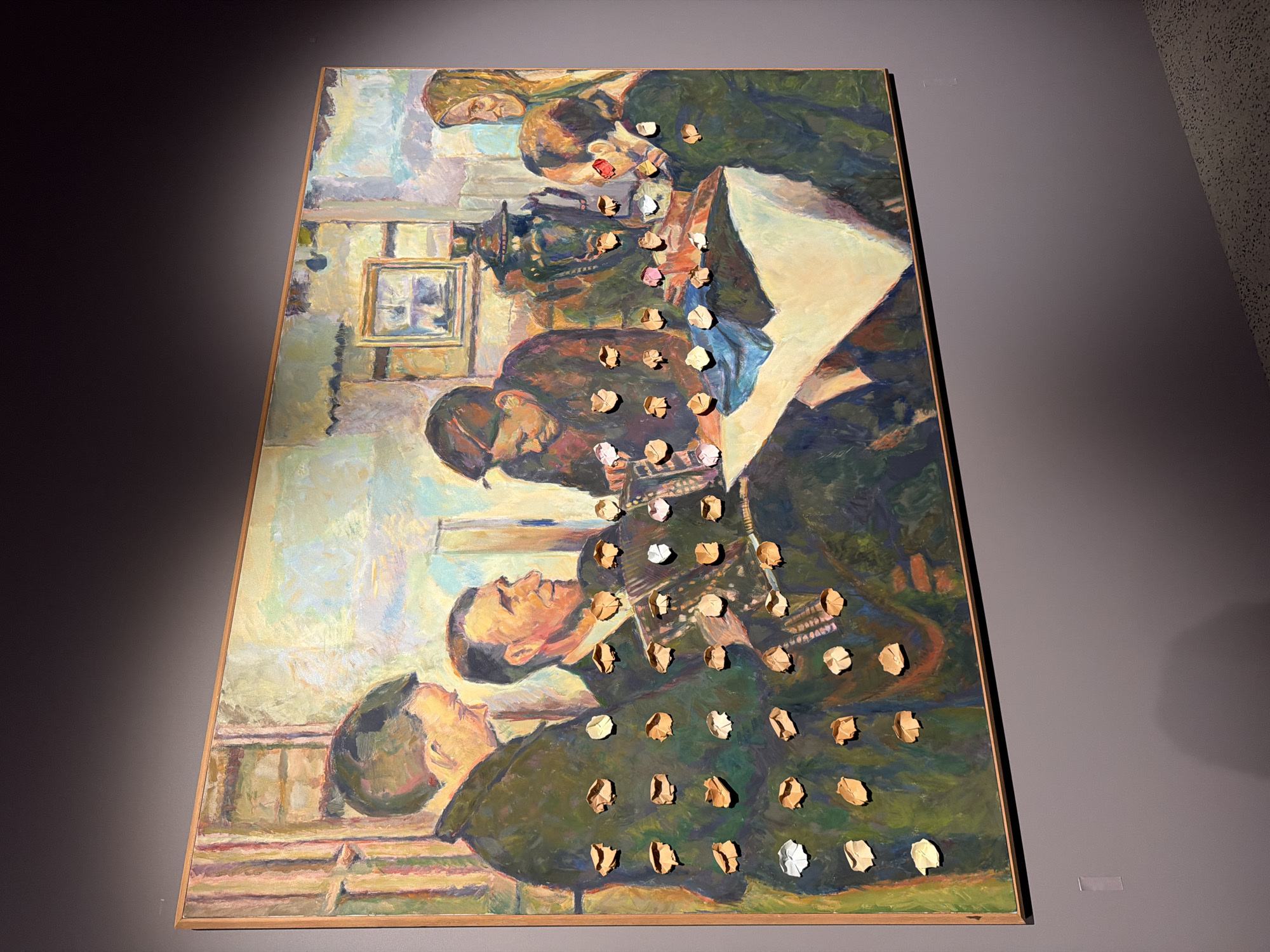 This is a photograph of a large, framed oil painting with a wooden frame. The painting depicts a group of people in what appears to be an indoor setting, possibly a studio or a room with a window. The style is impressionistic, with visible brushstrokes and a blend of colors, primarily greens, blues, and earth tones.
The subjects are rendered in a semi-abstract manner. Several figures are depicted in what seems to be a collaborative or perhaps a discussion-based activity. One figure, in the foreground on the left, is looking upwards and to the right. Another figure in the middle ground is bent over, seemingly focusing on something in front of them. Further back, two figures are shown interacting, one with their head tilted downwards. There are also suggestions of furniture or structures within the scene, including what might be a framed picture on the wall and elements resembling a table or benches.
A distinctive feature of the painting is the presence of small, three-dimensional, crumpled pieces of what appears to be paper or fabric adhered to the surface of the canvas in a grid-like pattern. These additions create texture and depth, breaking the flatness of the painted surface. The crumpled elements are in various shades of beige, white, and pink.
The lighting in the painting is soft, suggesting either an overcast day or an interior setting with diffused light. There are no clear indicators of the time of day or weather conditions outside. The overall emotion conveyed is one of focus and perhaps contemplation among the figures. No text is visible within the painting.