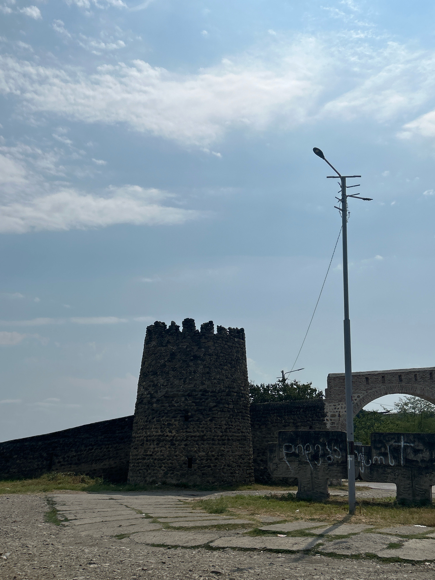 This is a full-shot, vertical photograph taken outdoors. The scene is a public space featuring historical architecture and a utility pole.
In the foreground, a rough, gravel path leads towards a paved area with large, cracked concrete slabs. To the left, a section of an old, dark stone wall runs diagonally, culminating in a rounded, crenellated tower made of similar dark stone. The tower has several small, rectangular openings and a crenellated top.
To the right of the tower, a tall, grey utility pole stands prominently. It has a single lamp and several horizontal arms with wires attached. A thin wire extends from the pole towards the left side of the frame, near the top of the tower.
Behind the utility pole and the tower, there is an arched stone structure, part of a larger building or wall, with a row of small, rectangular openings along its top. Some greenery is visible behind the structures. In front of the arched structure and to the right of the utility pole, two large, cross-shaped stone monuments stand. White graffiti is visible on the face of these monuments.
The sky above is a light blue with scattered white clouds. The lighting suggests it is daytime, possibly mid-morning or afternoon. The image appears to depict a historical site in the city of Vakiri, Georgia. There are no people visible in the image.