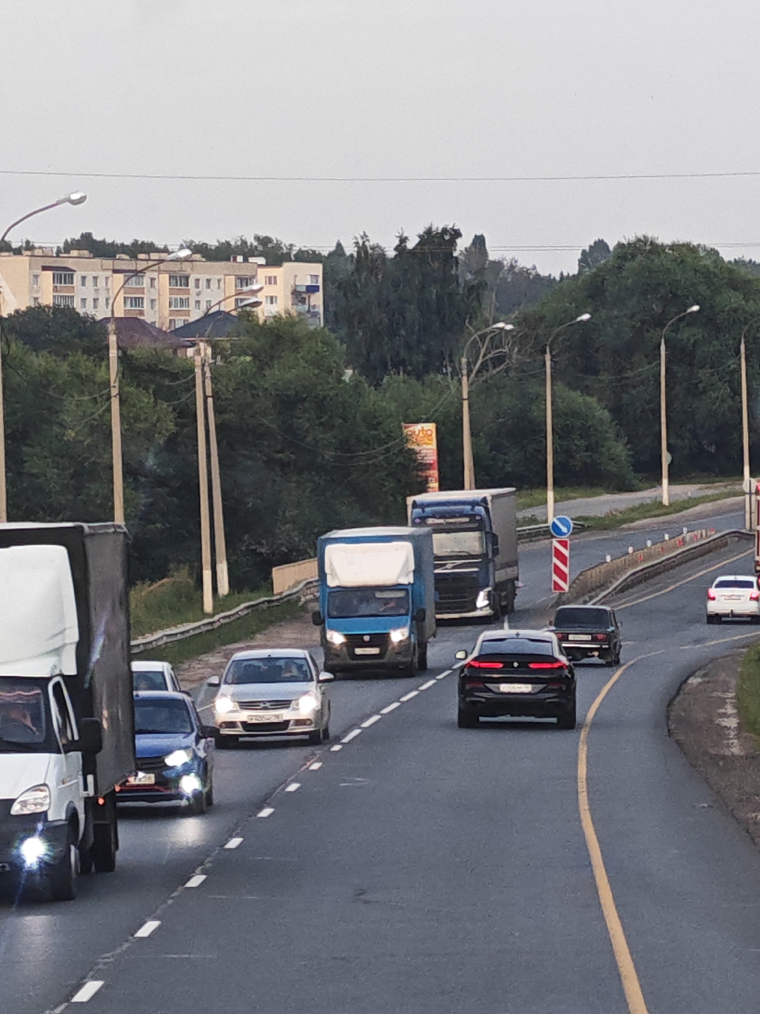 The image depicts a traffic scene on a highway. Several vehicles, including trucks and cars, are visible on the asphalt road. The environment is characterized by a cloudy sky and green trees lining the roadside. In the background, residential buildings and streetlights can be observed. The cars are in motion or stopped, indicating a traffic situation. The specific location is identified as Chemodanovka, Russia.