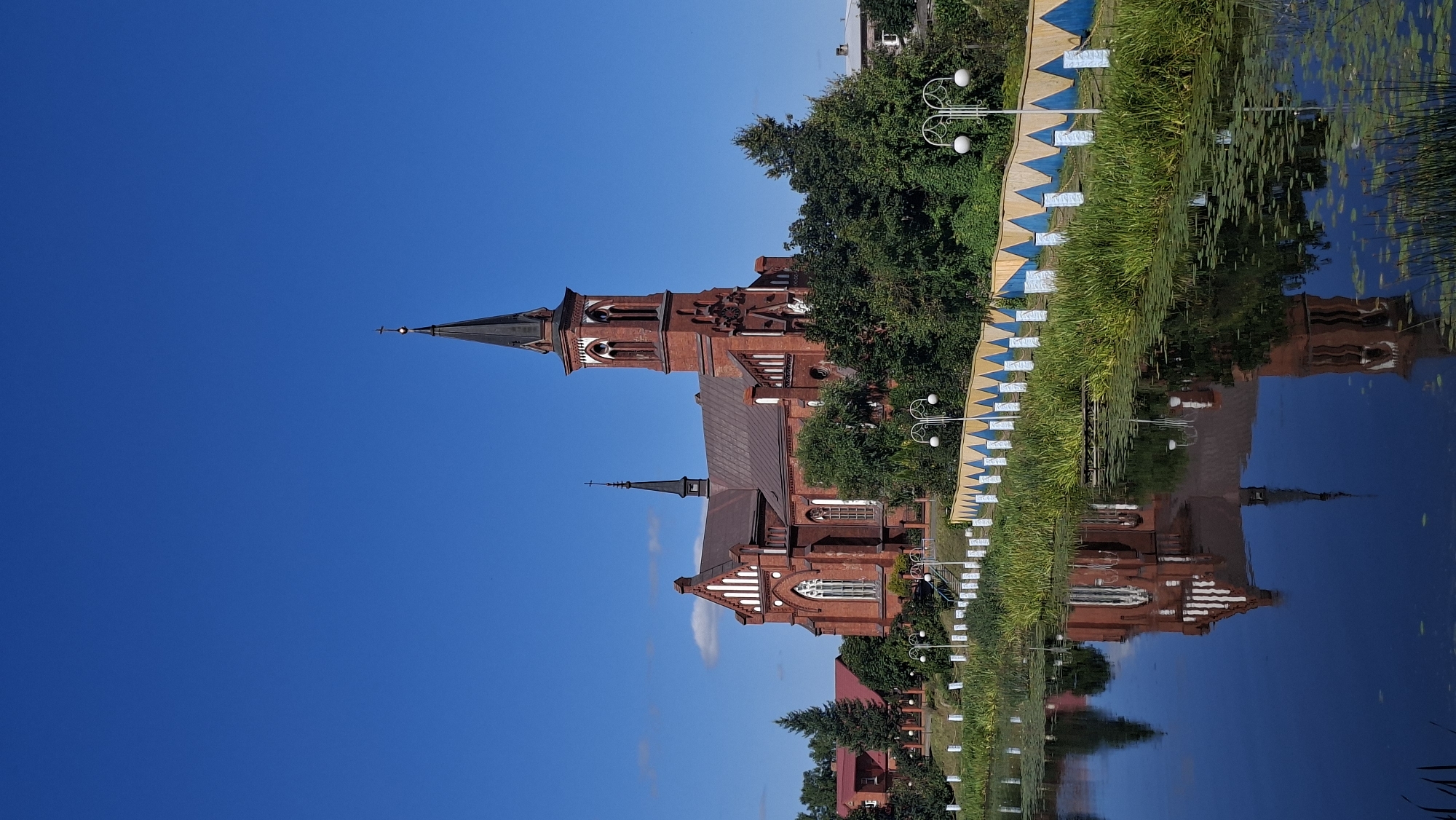 A brick church with a tall, slender spire dominates the right side of the image. The church is red brick with white trim around the arched windows and roofline. A smaller spire is visible behind the main one. The church is surrounded by lush green trees. In the foreground, a calm body of water reflects the church and the trees. Along the edge of the water is a walkway with a fence decorated with blue and yellow triangular patterns. White lampposts with globe-shaped lights are spaced along the walkway. The sky is a clear, vibrant blue. The scene appears to be the Church of the Transfiguration of Jesus in Pastavy, Belarus, given the architectural style and its characteristic location next to a body of water.