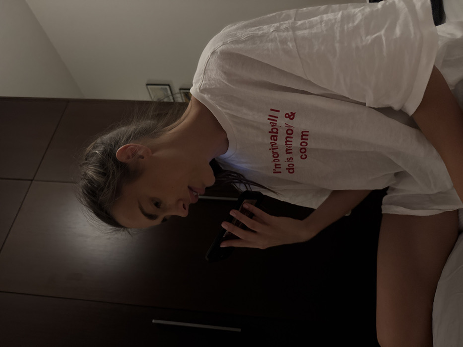 A woman is seen in a close-up shot, looking down at her phone. She is wearing a white t-shirt with red text printed on it. She is positioned in front of a dark brown, possibly wooden, wardrobe or storage unit. The image has a casual, candid feel, possibly taken indoors in a bedroom setting, suggested by the edge of a bed in the corner of the frame. The lighting seems to be soft and diffused.