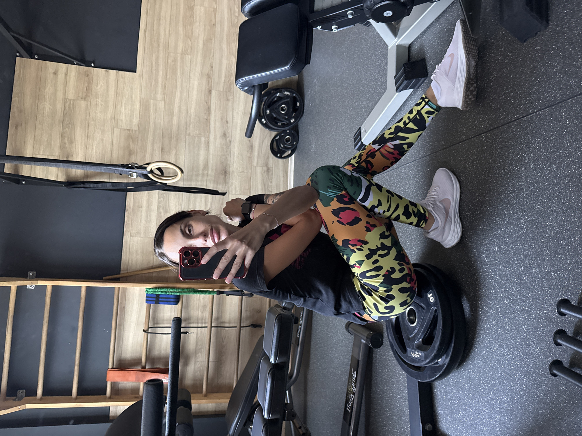 A young woman is in a gym, sitting on a weight plate on a weight machine. She is holding a phone and facing the camera, apparently taking a selfie. She is wearing a black t-shirt, patterned leggings, and light-colored sneakers. The gym has a gray floor and a wooden wall in the background with a climbing frame. Other exercise equipment is visible, including a leg press machine and other weight plates. The location is Pleven, Bulgaria.
