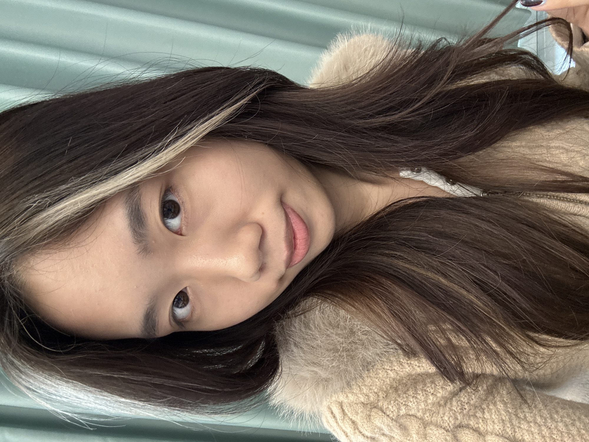 A young adult female of East Asian descent is depicted in a close-up, chest-up portrait taken indoors, likely in Nan'an District, China. She possesses long, dark brown hair with a distinct, lighter-colored highlight streak on the viewer's left side of her head. Her dark eyes maintain direct contact with the camera, and her lips are subtly parted in a slight, closed-mouth expression. She is dressed in a beige-colored knitted sweater featuring a cable-knit pattern on the shoulder and a light brown faux fur collar. A white garment with a zipper and metallic fasteners is visible at her neckline beneath the sweater. The lower right portion of the image shows her left hand, adorned with dark-colored nail polish, gently holding a strand of her hair. The background consists of a uniformly light teal or mint green pleated curtain.
