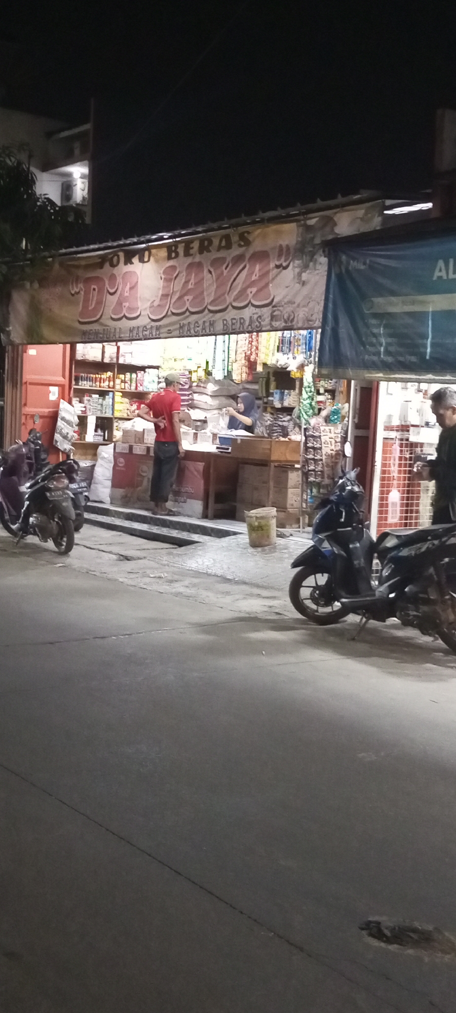 The image depicts a street-level view of a shop in Setu, Indonesia, at night. The shop, identified by a banner reading "TOKO BERAS 'DA JAYA'" (Rice Shop 'Da Jaya') and a smaller sign "MENJUAL MACAM - MACAM BERAS" (Selling Various Kinds of Rice), is illuminated and displays shelves stocked with various goods. In front of the shop, a man wearing a red shirt and a cap stands at a counter, interacting with a woman wearing a hijab who is behind the counter. Two motorcycles are parked in front of the shop, one on the left and another on the right. A third motorcycle is visible parked further down the street on the right. The street surface is paved, with some visible cracks and debris. The overall environment suggests a commercial area operating into the evening hours.
