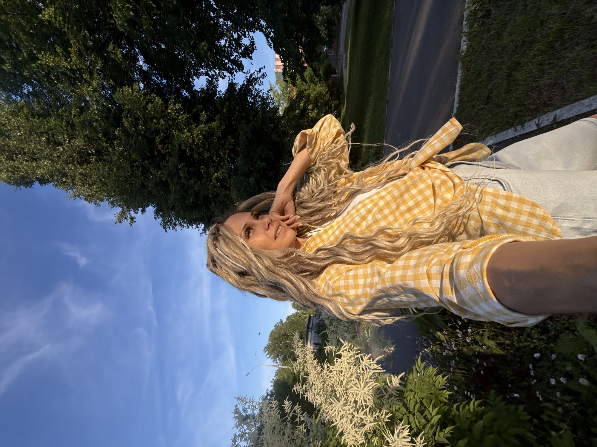 In Заволжье, Russia, a young woman with blonde dreadlocks and a yellow checkered shirt is captured in a selfie outdoors. She is standing near a lush green area with trees and white flowering bushes. The sky is a clear blue with a few wispy clouds, indicating it is daytime. The woman is leaning her head on her hand, looking to the side with a slight smile, suggesting a relaxed and happy mood. The background shows a paved path and a grassy area, with buildings visible in the distance. The lighting suggests it is late afternoon or early evening, with the sun casting a warm glow on the scene.