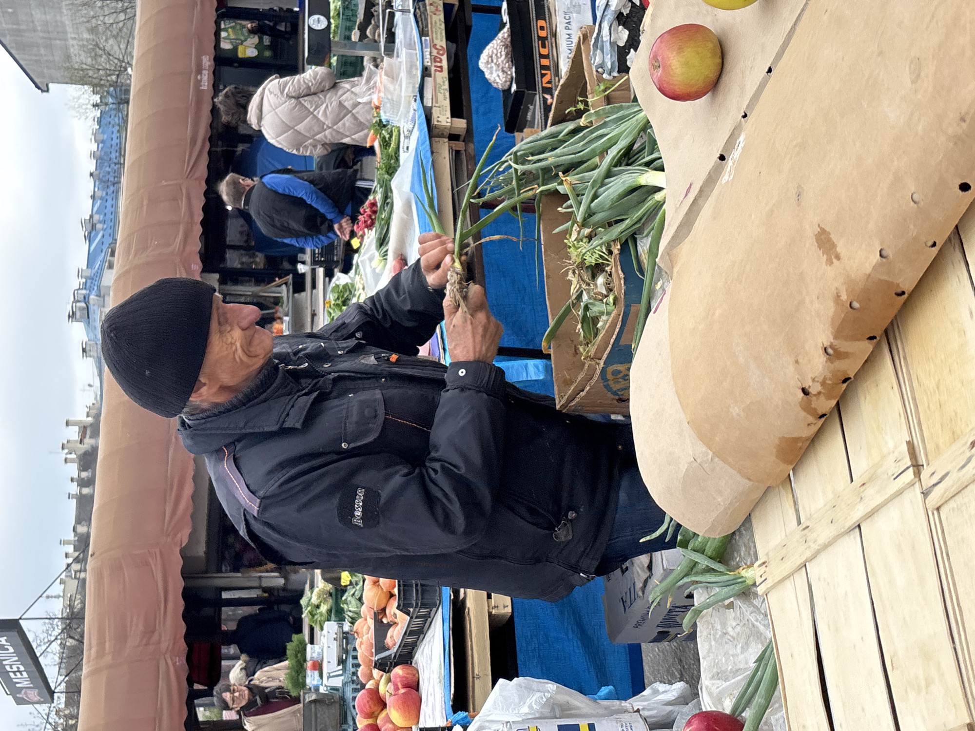 An eye-level, tilted, outdoor shot at a Zagreb, Croatia market shows an older man in a black beanie and jacket preparing green onions for sale. He stands behind a wooden table covered in various produce, with other vendors and customers in the background. He is actively cleaning or trimming a bunch of green onions. The market has a light-colored awning. The sky is overcast.