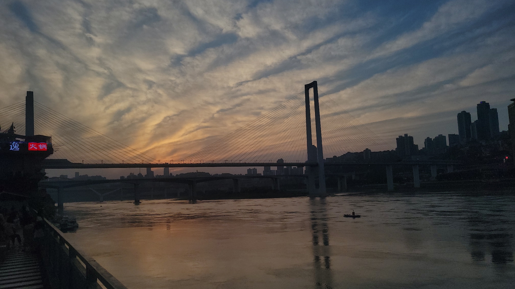 The image presents an urban river landscape at sunset or twilight, characterized by a vibrant sky, multiple bridges, and a city skyline. The sky features a gradient of colors, transitioning from warm oranges and yellows near the horizon to cooler blues and grays higher up, with streaky, elongated clouds across the frame.
Two prominent bridges span the wide, reflective river. The larger, cable-stayed bridge dominates the mid-ground, featuring two distinct pylons: one a single tall pillar on the left and the other an inverted U-shaped structure on the right. Numerous cables radiate from these pylons, supporting the bridge deck. Below it, a second, lower multi-span concrete bridge runs parallel across the river.
The river itself reflects the sky's warm hues, appearing as a shimmering expanse of orange and brown tones. A small, flat-bottomed boat with a single figure standing onboard is visible in the middle of the river, silhouetted against the water's surface.
On the far left, a multi-level structure or building is nestled on the riverbank, adorned with bright red neon signs. The Chinese characters "逸" (Yì) and "火锅" (Huǒguō) are clearly visible, indicating a hot pot restaurant or similar establishment. A few indistinct figures can be seen on stairs or a walkway in the lower-left foreground. The opposite bank features a dense skyline of high-rise buildings, largely silhouetted against the fading light, suggesting a bustling city.
The overall impression is one of serene urban beauty, capturing the transition from day to night over a major waterway.