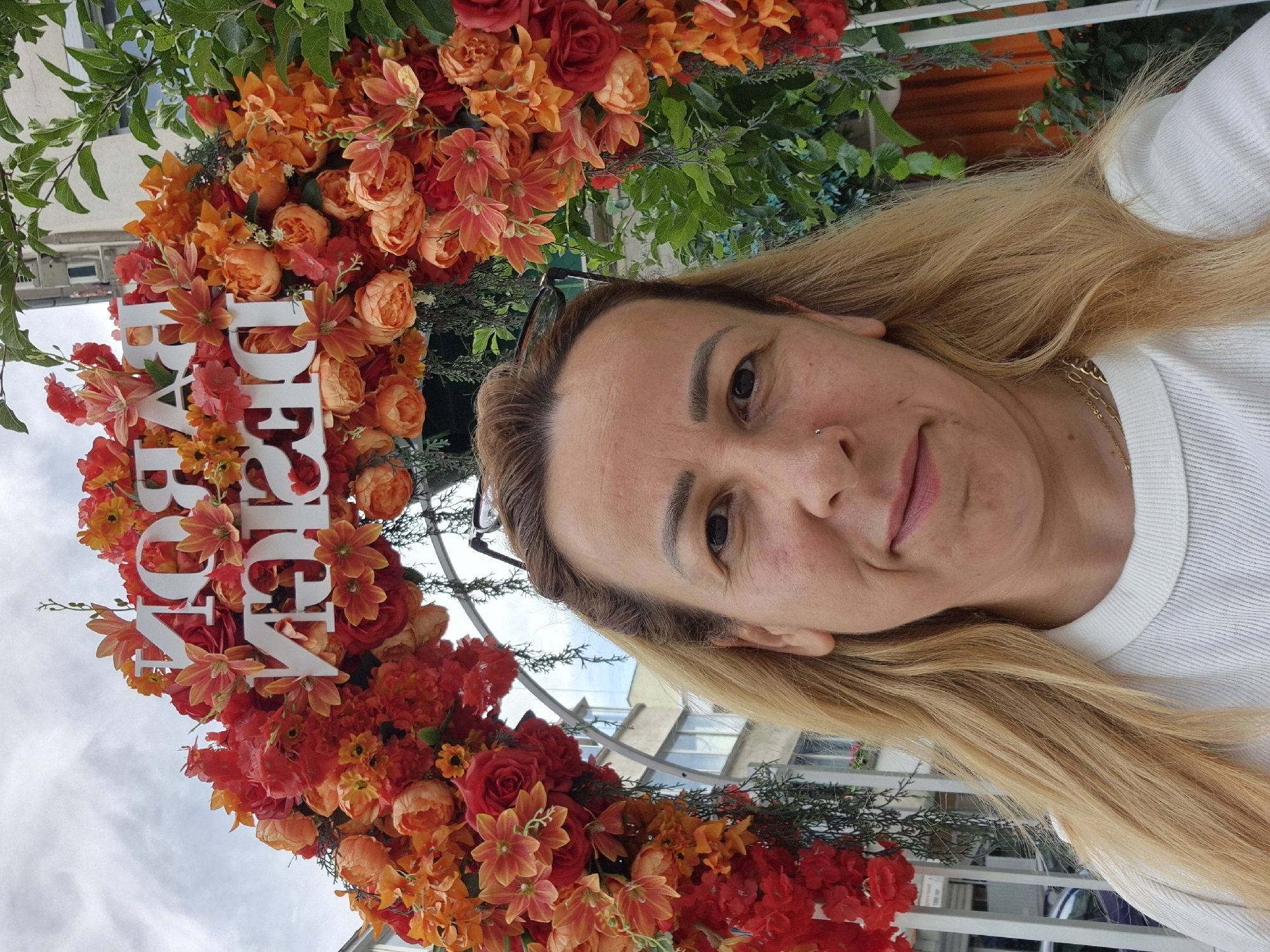 An outdoor, eye-level, medium shot shows a woman in the foreground with a floral archway behind her in the city of Unknown, Unknown. The woman has light brown hair and is looking at the camera. She is wearing a white crew-neck shirt. The floral archway is decorated with red, orange, and pink flowers. The words "PAROK DESIGN" are spelled out in white, three-dimensional letters across the archway. Behind the archway, there are buildings and trees. The sky is cloudy.