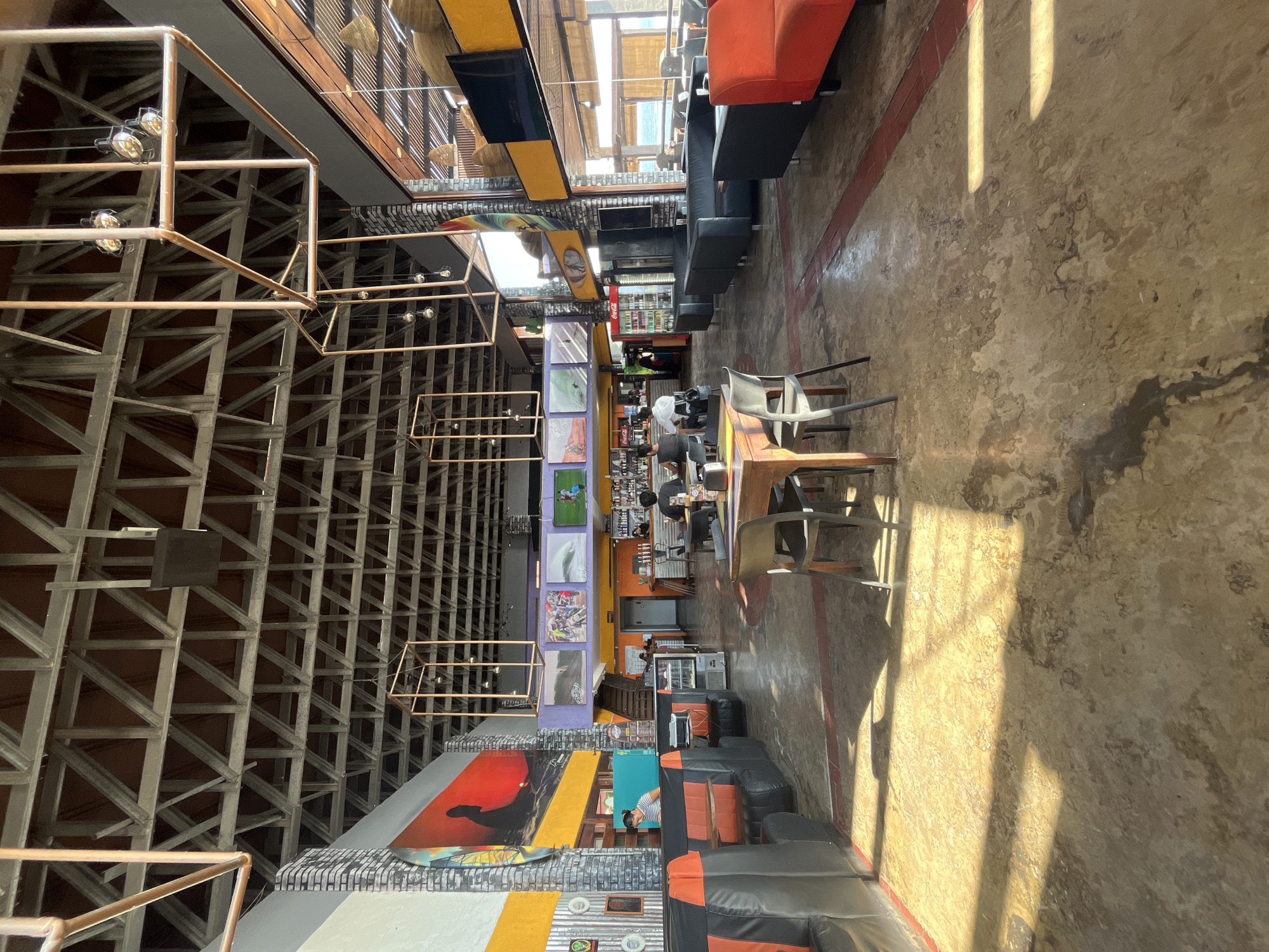 The image shows the interior of a restaurant or café in Arica, Chile. The layout features tables and chairs for dining, with booth-style seating along the right-hand wall, some of which is orange and black. The floor has a textured concrete finish with streaks of color. The left side of the image is dominated by a complex framework of metal beams, above a vertical display of surf-themed artwork and advertising. People are seated at tables in the center of the room, suggesting a partially occupied space. The background shows a bar area and further seating, as well as glimpses of the outside world. Natural light pours in from above, illuminating the interior.