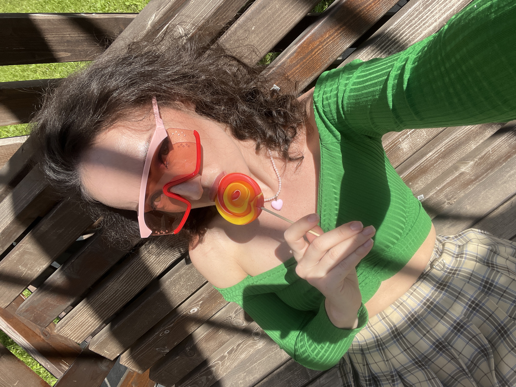 A person is lying on a wooden bench outdoors, with a lollipop in their mouth and a pink-framed pair of sunglasses on. They are wearing a green off-the-shoulder top and a plaid skirt. Sunlight casts shadows on the bench and the person. The background shows green grass. The scene is set in Kazan, Russia.