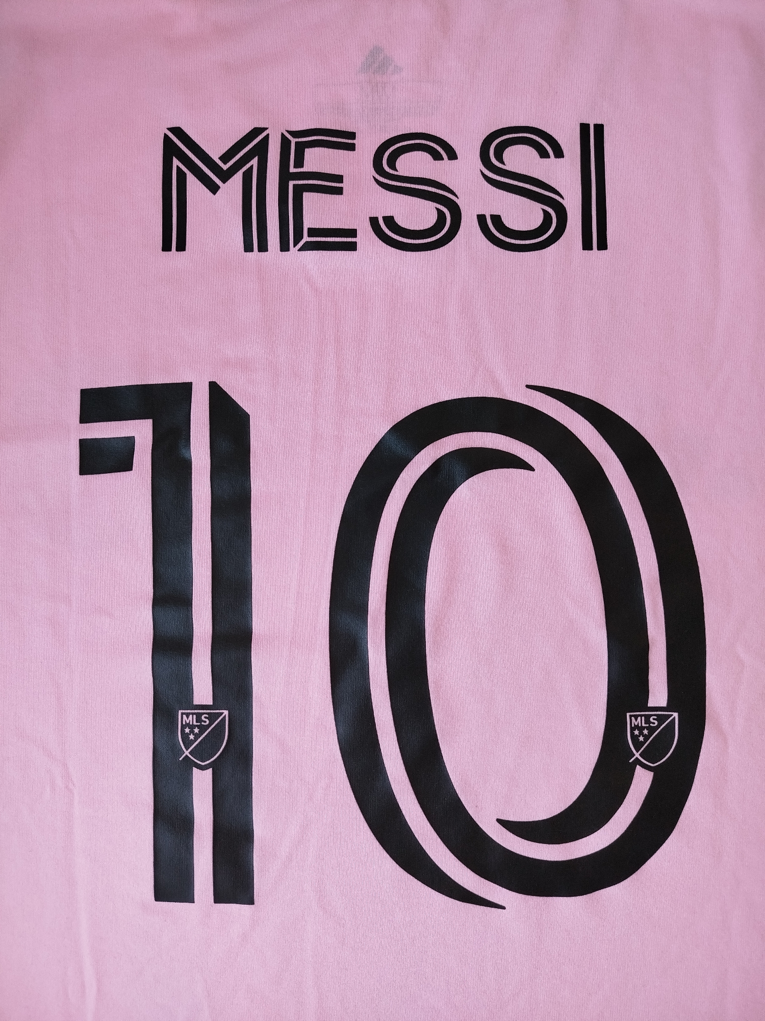 The image shows the back of a pink soccer jersey. In black lettering, the name "MESSI" is printed across the upper portion of the jersey. Below the name, the number "10" is printed in black, with the MLS logo visible within the bottom of the "1" and the "0". The overall image is a close-up shot of the jersey. There is no information about the city of Varna, Bulgaria in the image.