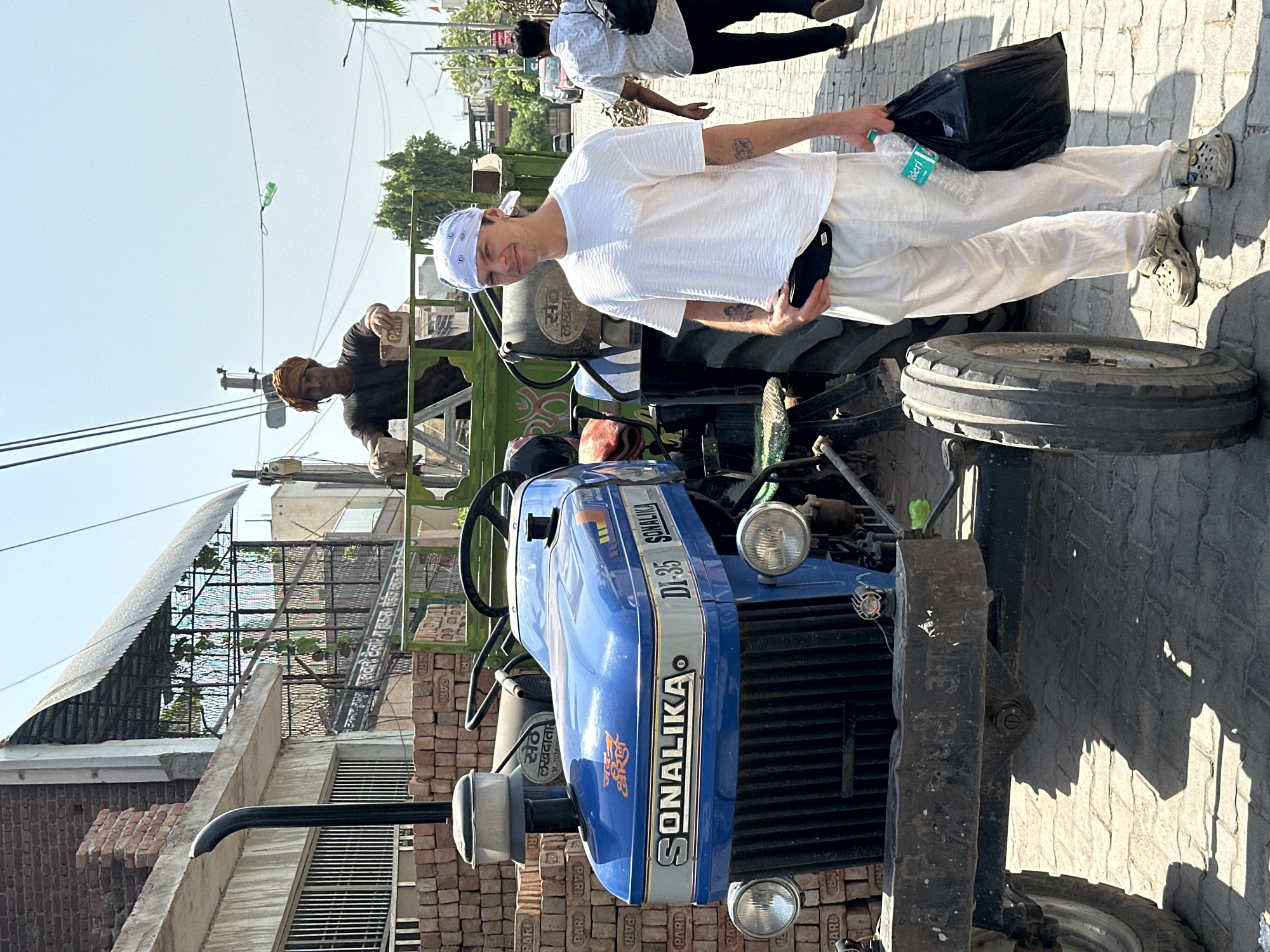A man with a bandana poses next to a blue Sonalika DI-35 tractor on a brick-paved street in Mathura, India. The tractor is parked in front of stacked bricks. Another man is sitting on the tractor behind him. A man is walking on the side of the street with a black bag and a water bottle.