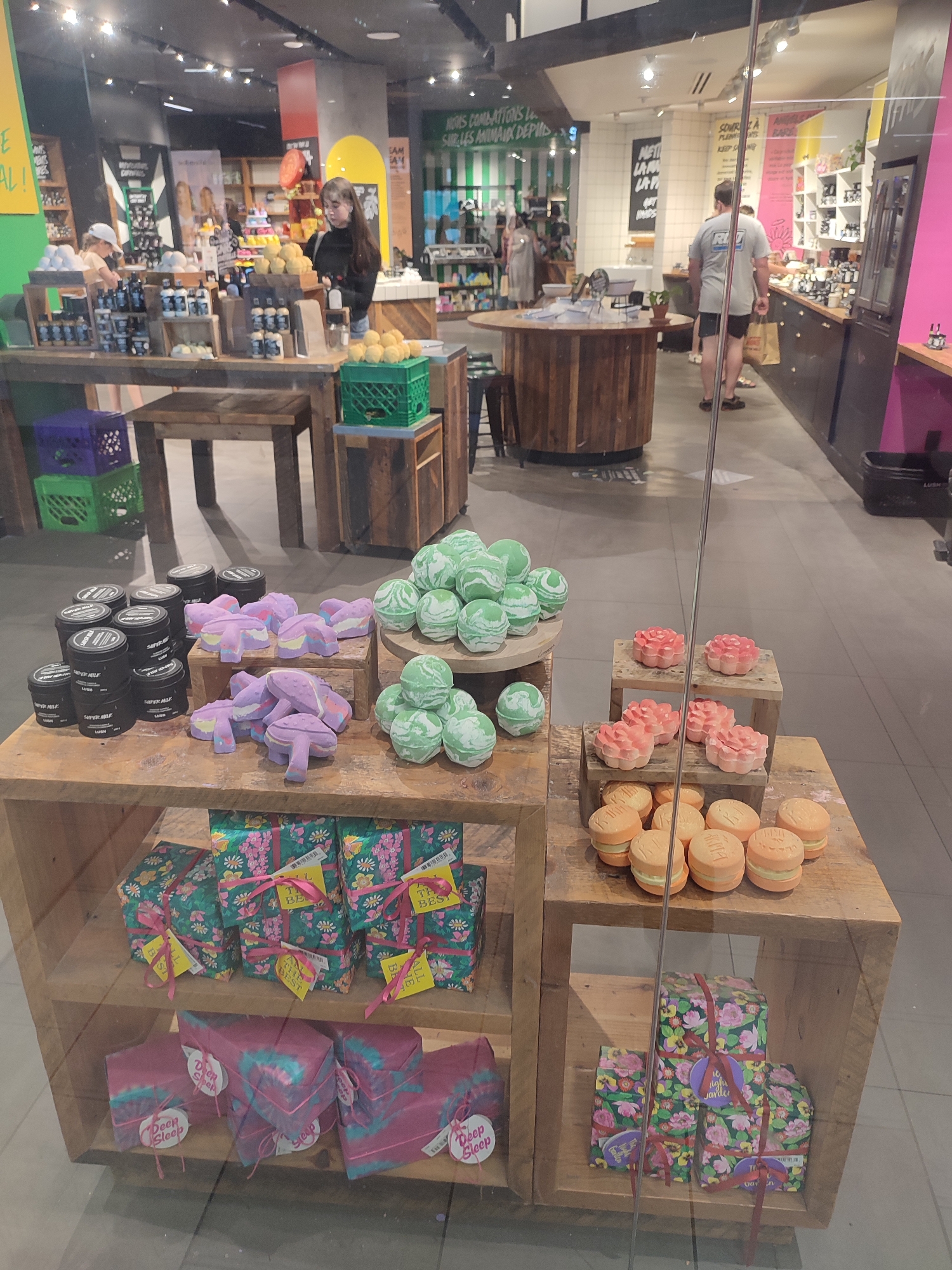 This is a medium shot image taken through a glass window of a retail store, likely LUSH Cosmetics, in Montreal, Canada. The store appears brightly lit, suggesting it's daytime. The scene showcases an array of bath bombs and gift sets arranged on wooden shelves and displays.
In the background, several people are visible. Two women are in the distance, one with dark hair in a black top and a young girl wearing a white cap and a light-colored shirt, both appearing to be staff or customers browsing. Another man in a grey t-shirt and black shorts is also seen, seemingly interacting with a display. The store interior features multiple shelves filled with products, promotional signage, and decorative elements like a large wooden circular table.
The foreground is dominated by product displays. On the left, stacks of black cylindrical containers, possibly skincare products, are arranged. To their right, purple, mushroom-shaped bath bombs are displayed on tiered stands. Further right, numerous green and white swirled bath bombs are grouped on round platters. Below these, gift-wrapped boxes with floral patterns and pink ribbons are stacked. On the bottom shelf, more gift boxes, some with a tie-dye effect in pink and teal, are visible. In the far right foreground, an assortment of round, orange bath bombs, resembling mini burgers, are displayed on a wooden shelf, with pink, flower-shaped bath bombs above them.
Visible text includes "SUPER MILK" on the black containers, and tags on the gift boxes read "TH BEST" and "ALL THE BEST." On the pink and teal tie-dye wrapped gifts, the text "Deep Sleep" is clearly legible. Other fragmented text, possibly store slogans or product names, is present but not fully decipherable. The overall impression is one of a vibrant, organized retail environment showcasing a variety of colorful bath products.