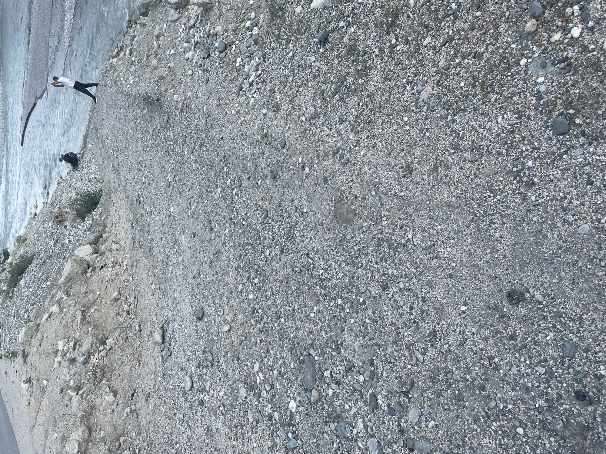The image depicts a rocky, barren landscape adjacent to a body of water, likely a river, in Khodzha-Obigarm, Tajikistan. The foreground is dominated by a gravelly path leading towards the water. In the mid-ground, two individuals are visible near the water's edge. One person is crouched down, while the other stands, appearing to be taking a photograph with a mobile phone. The background shows the expansive river flowing between rocky banks. The overall environment is dry and undeveloped, characteristic of a natural, outdoor setting.