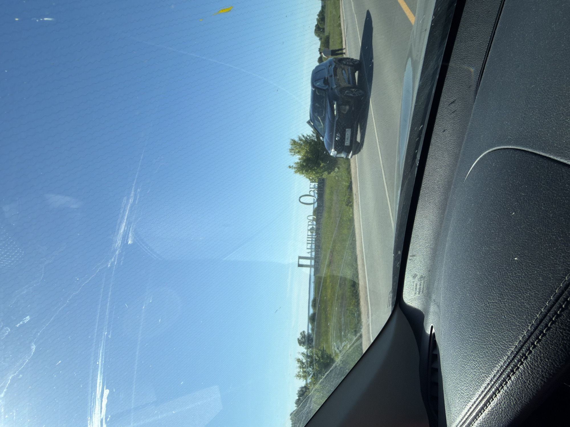 The image depicts a daytime scene captured from inside a vehicle, looking forward through a windshield. The vehicle's dark, textured dashboard with visible stitching on the left is prominent in the foreground. The windshield exhibits multiple streaks, smudges, and reflections, as well as a faint hexagonal or wavy grid pattern.
In the mid-ground, a two-lane asphalt road is visible. A dark-colored Hyundai Santa Fe SUV, appearing to be a 4th generation model, is positioned on the right shoulder of the road, facing towards the camera. The vehicle's Russian license plate "P744OX 37" is partially discernible, indicating the Ivanovo Oblast region of Russia. A single individual, dressed in a light-colored top and dark trousers, stands near the rear passenger side of the Hyundai SUV on the road's shoulder.
Further in the mid-ground, to the left of the road, a large, free-standing sign with white Cyrillic letters reads "ПЛЕЩЕЕВО ОЗЕРО" (Pleshcheyevo Lake). This sign is situated on a grassy embankment. Beyond the road and sign, the environment consists of a green landscape with scattered trees and bushes. In the distant background, a large body of calm water, consistent with a lake, is visible under a clear, bright blue sky. The sunlight is strong, creating visible reflections on the windshield.
The identified location is near Lake Pleshcheyevo, likely within the Pereslavl-Zalessky District, Yaroslavl Oblast, Russia.