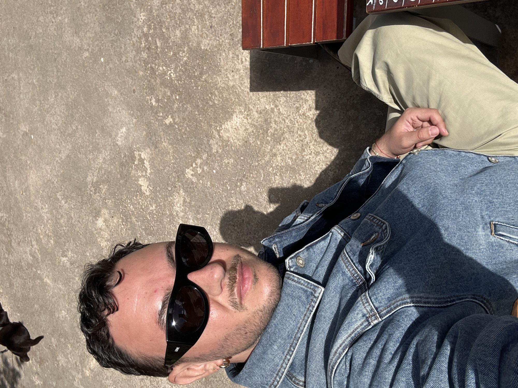 A medium-shot captures a person wearing black sunglasses and a denim jacket, seated beside a wooden bench with beige pants. The person is positioned on the right side of the frame. In the lower left corner, a small black dog is visible. The scene is set outdoors on a concrete surface, possibly a sidewalk or patio. Although the location is not directly identifiable as Madrid, Spain from the visual details, the person appears to be enjoying a sunny day in an urban environment.