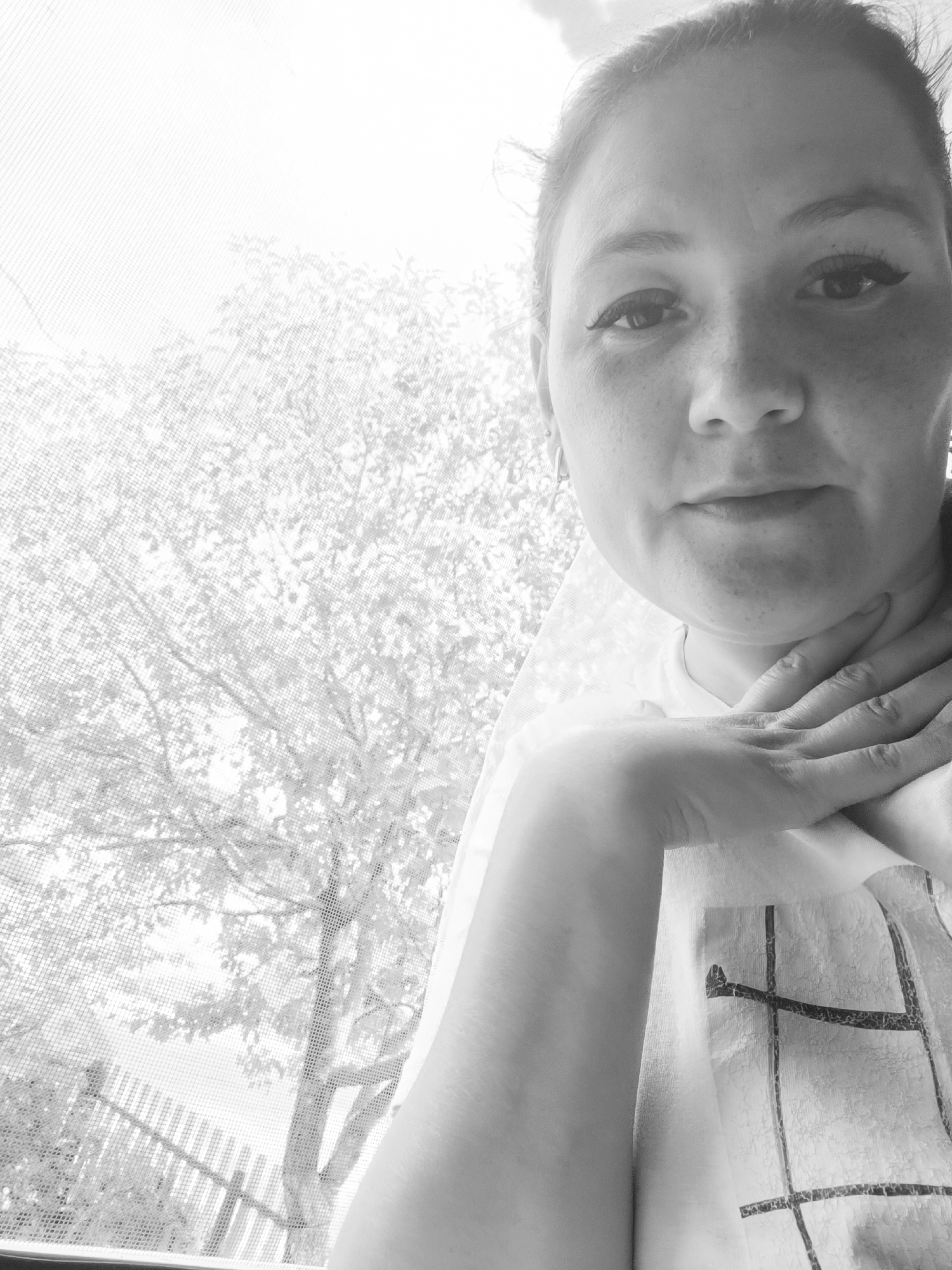 The black and white image shows a close-up of a woman looking at the camera, with a window screen and foliage in the background. The woman has dark hair pulled back and is wearing a t-shirt with a graphic design. Her left hand is placed on her neck. The scene appears to be indoors, near a window, with a tree and a fence visible outside through the screen. There is no identifiable information in the image that would indicate the location is Karsha, Russia.
