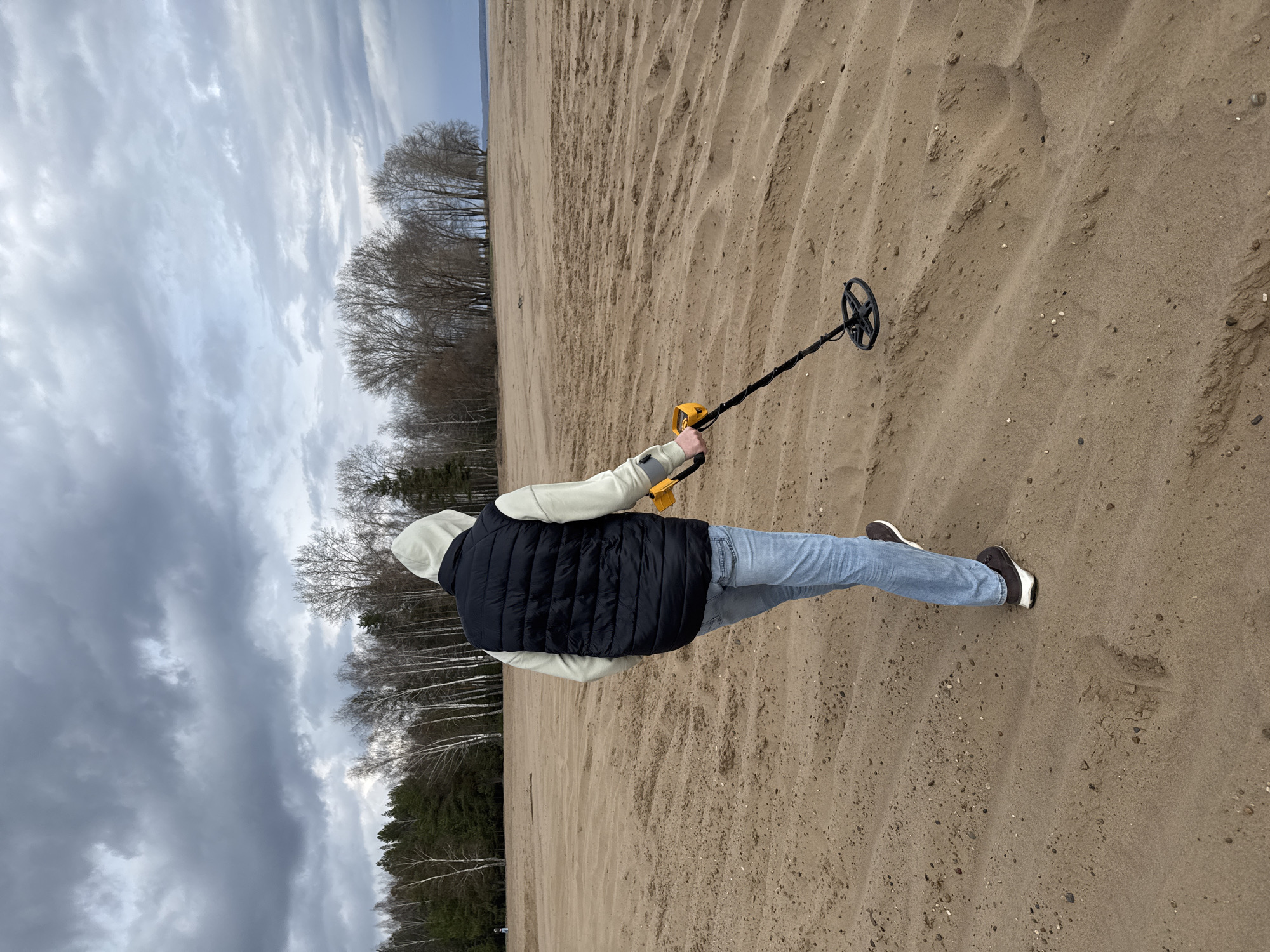 A person stands on a sandy beach, metal detecting. They face away from the camera, holding a yellow metal detector with a black coil. The person is wearing a white hooded sweatshirt, a black vest, blue jeans, and black shoes. The beach consists of sand with ridged patterns. In the background, there is a treeline and a cloudy sky. The location is likely in Chaikovsky, Russia, based on available information about similar landscapes in the area.