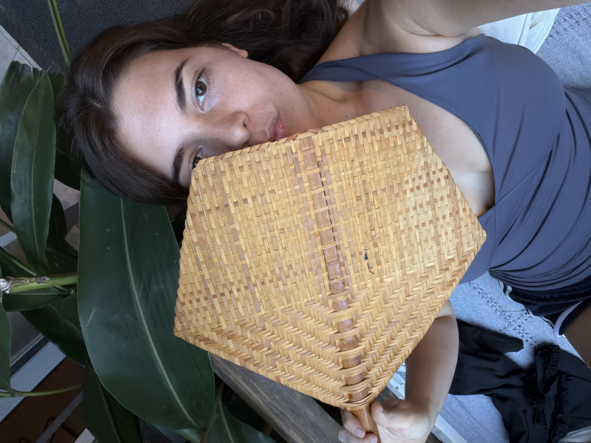 The image depicts a woman with brown hair and light skin lying down. She is wearing a blue tank top. Her head rests near a large green leaf, possibly from a plant. She is holding a woven hand fan in front of her face. The background suggests an interior setting with a glimpse of a gray or neutral-toned surface, possibly a couch or bed. The image has no explicit indicators connecting it to Vicente López, Argentina.