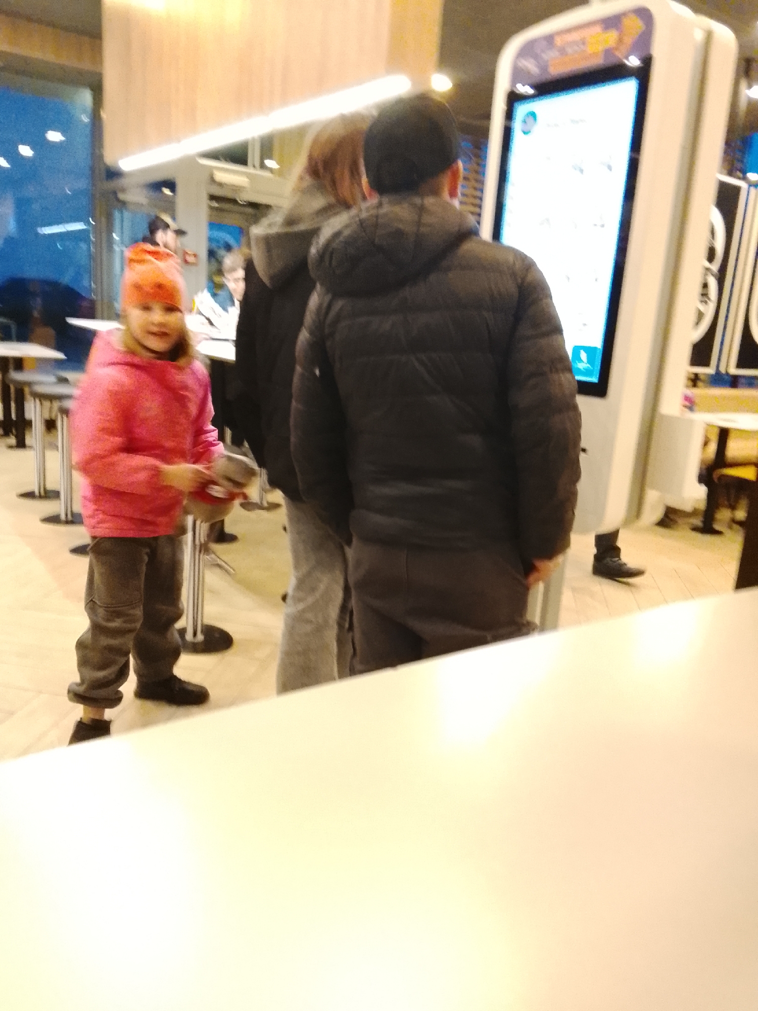 The image depicts a scene in what appears to be a McDonald's restaurant in Obninsk, Russia. A young girl wearing a pink jacket and orange hat stands with a stuffed animal. Next to her, two individuals are using a touchscreen kiosk. The environment includes tables and chairs, with large windows showing a view of the city outside. Additional patrons are visible in the background. The overall lighting is bright and fluorescent.
