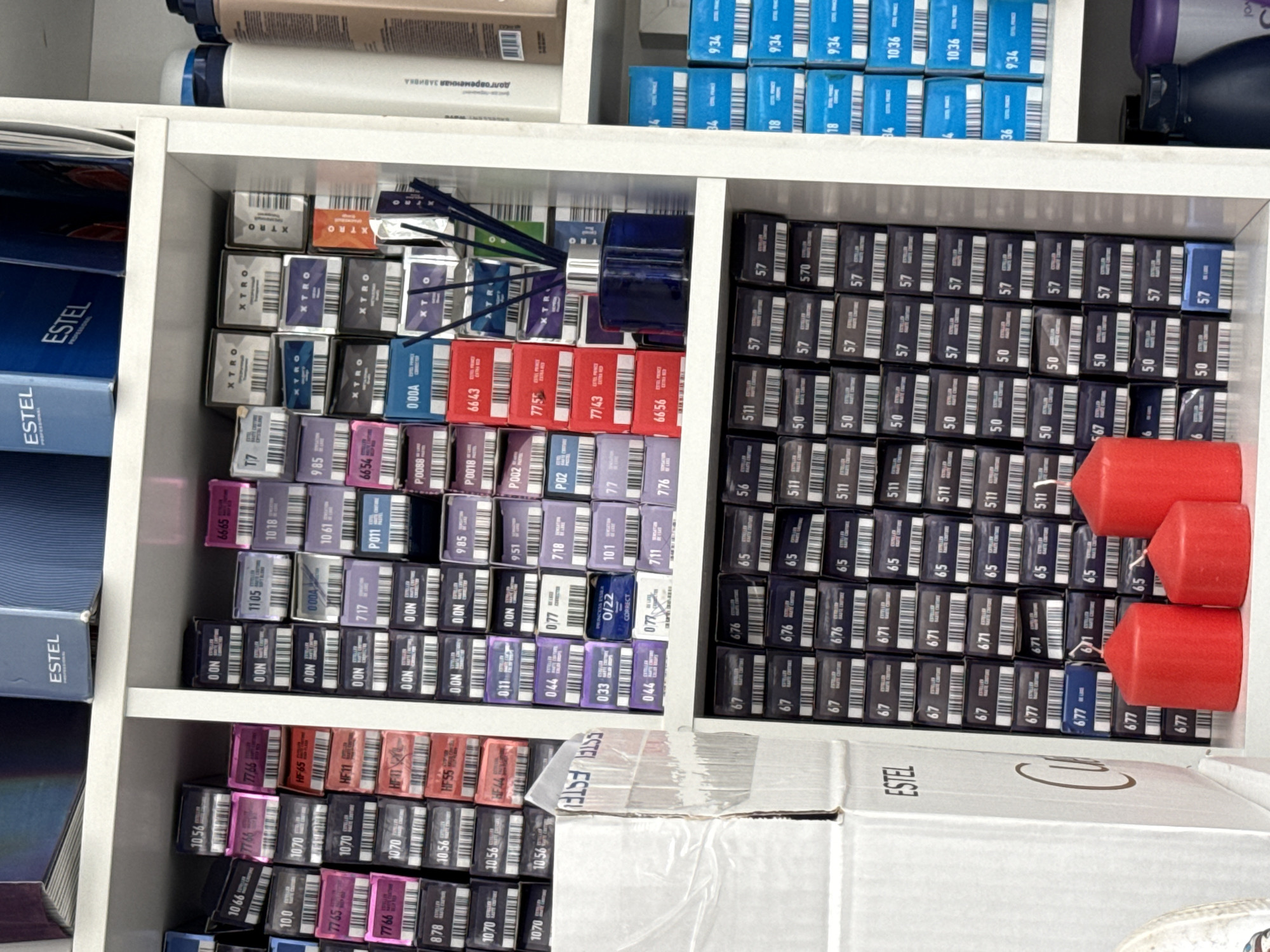 The image shows a shelf in a retail store, likely a hair salon or beauty supply store, displaying various hair dye products and other beauty items. The products are arranged in rows and columns on white shelves. The hair dye boxes are predominantly black and grey with colorful labels indicating different shades. Some of the prominent brands visible are "ESTEL" and "XTRA." In the foreground, on the bottom shelf, are three red candles. To the right of the hair dye shelves, there are several tall bottles of hair products, some white and some colored. A blue diffuser with reeds is placed on a shelf in the middle section. The store appears to be well-organized with a clean and bright environment. There are no people visible in the image. The presence of the "ESTEL" brand, a prominent Russian professional cosmetics manufacturer, suggests the location is likely within Russia. Based on the product branding and typical retail setups for such products, this scene is consistent with a beauty supply store or a professional salon in Chaikovsky, Russia.