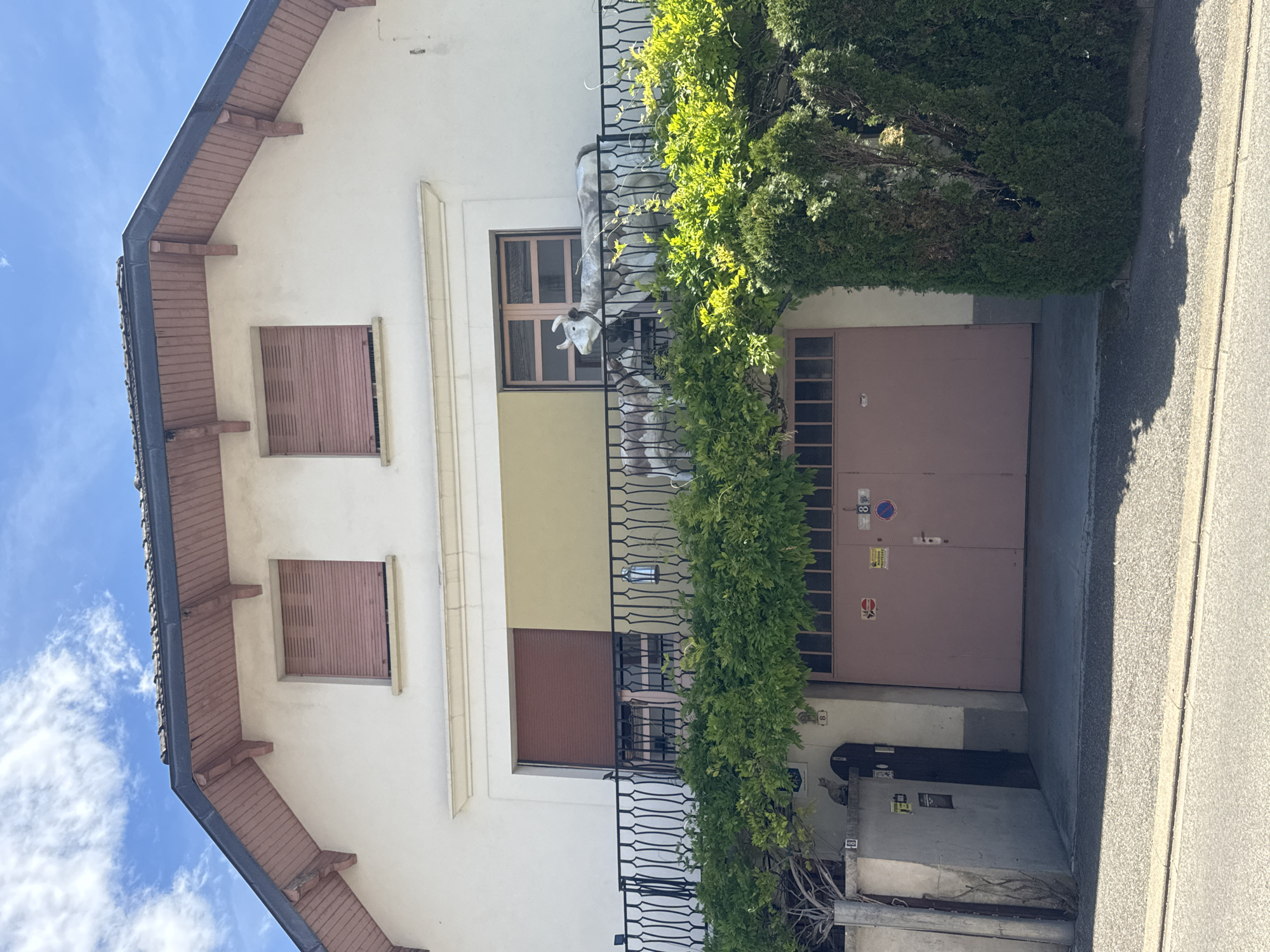 This is a full shot, eye-level, outdoor photograph taken on a bright, sunny day. The image features a two-story house with a light-colored facade, a dark-tiled roof, and a prominent wooden soffit painted a reddish-brown. Two identical windows with closed, beige shutters are visible on the upper level. Below these, a balcony with a black wrought-iron railing spans the width of the house. A yellow-painted section is visible behind the balcony. Two decorative cow statues are placed on the balcony, facing outwards. A lush green vine with vibrant leaves covers the railing, extending downwards.
In front of the house, there's a large, light-pink double garage door with a horizontal paneled design at the top. Several stickers and signs are affixed to the garage door and the wall to its left. To the left of the garage, a lighter-colored wall section is visible, and a dark wooden door is set into the wall. A gray cat is perched on top of this wall section, to the left of the wooden door.
The environment appears to be a residential area. The house is situated on a slight incline, with a paved driveway leading to the garage. A paved sidewalk runs in front of the house, separated from the property by a curb. Greenery, including bushes and trees, surrounds the house, contributing to a somewhat rustic aesthetic. The sky above is bright blue with scattered white clouds. The photograph is sharp and well-lit, capturing the details of the architecture and the surrounding environment. The scene is located in Coteaux du Lizon, France.