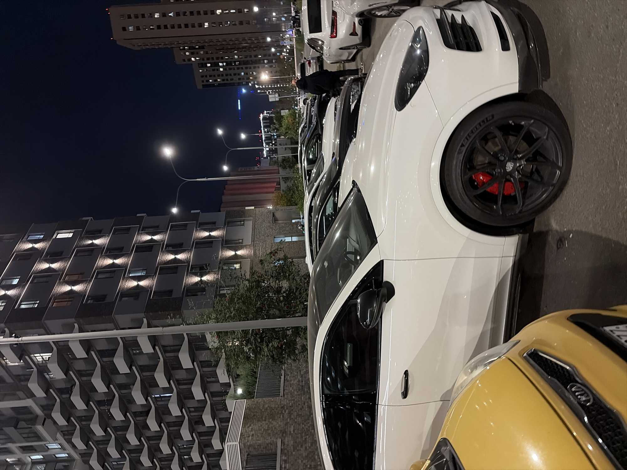 At night, a row of parked cars, including a white Porsche Cayenne with red brake calipers, a yellow Kia, and several other vehicles, are visible in front of a modern, multi-story residential building in Moscow, Russia. The building features a facade with numerous balconies, illuminated by outdoor lighting. In the background, other tall buildings with lit windows and streetlights line the urban landscape. A lone individual, wearing a dark hooded sweatshirt, stands between two cars, facing away from the camera. The scene depicts a residential area at night, with the focus on the parked cars and the contemporary architecture of the surrounding buildings.