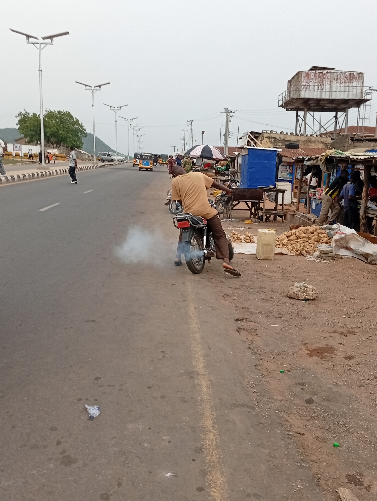 A man on a smoking motorcycle is parked on the side of a road in Zango-ndaji, Nigeria. He wears a tan shirt and brown pants, and is facing away from the camera. To the right of him are piles of potatoes for sale next to makeshift stalls. In the background, there's a water tower, a blue kiosk, and several people standing around. The road is asphalt, and to the left, there's a pedestrian crossing. Further down the road, there are auto rickshaws and other vehicles. Street lights line the road, and there's a hill in the far distance. A small piece of trash is on the road in the foreground.