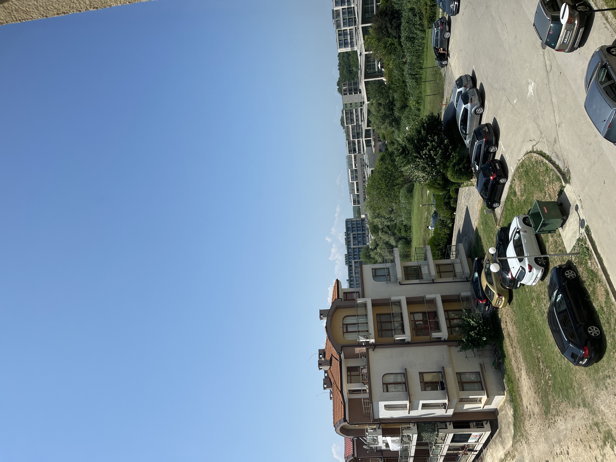 The image captures a bright, sunny day in the city of Obzor, Bulgaria, with a clear blue sky dominating the upper portion of the frame. In the foreground, a multi-story apartment building with a distinctive orange-tiled roof is prominently featured. The building has cream-colored walls and balconies with glass railings. Adjacent to the building is a parking lot with several cars parked.
To the left of the apartment building, a patch of dry, brown grass is visible. The parking lot itself is paved with concrete and shows some signs of wear. Several vehicles are parked in rows, including a black station wagon, a white hatchback, a gold sedan, a black sedan, and a silver SUV. A large green trash bin is also present in the parking area.
In the background, a row of modern, multi-story buildings with flat roofs stretches across the landscape, interspersed with lush green trees and vegetation. The overall impression is of a residential area, likely a resort town given the presence of multiple apartment complexes and the clear, sunny weather. There are no prominent signs or landmarks that specifically identify the location within Obzor, but the architecture and landscape are consistent with coastal resort areas in Bulgaria.