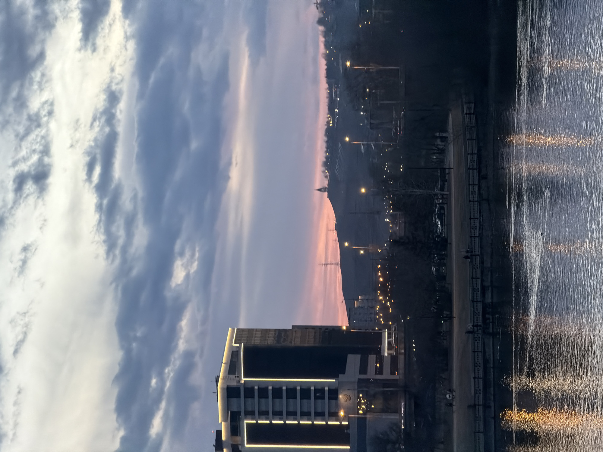 The image captures a cityscape at dawn or dusk. In the foreground is a modern building with horizontal light strips. Above and behind the building, the sky transitions from cloudy grey to pale pink, suggestive of sunrise or sunset. A hill is visible in the background, lined with streetlights. The presence of a water body is indicated by reflections of light on its surface, on the right side of the image. The location is in the city of Krasnoyarsk, Russia.
